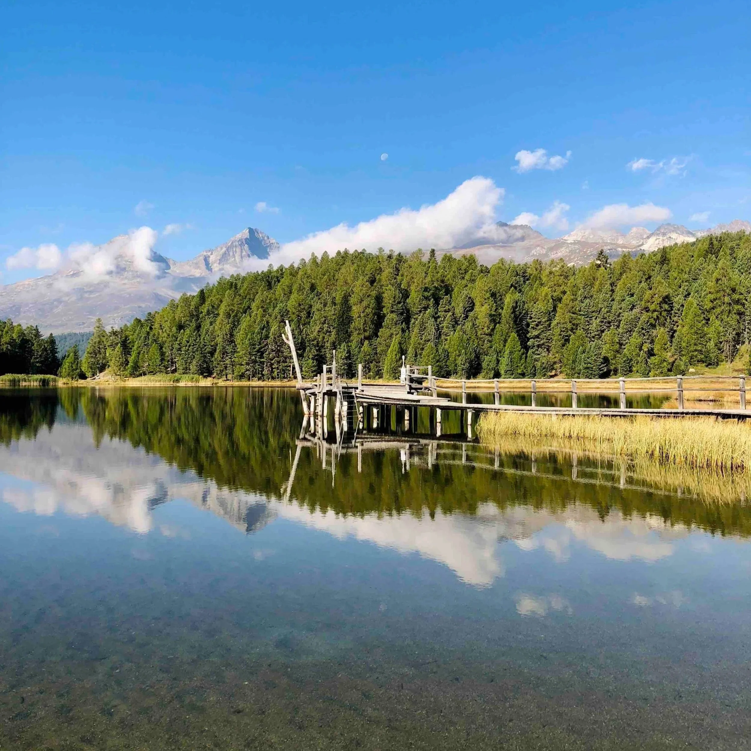 A serene mountain landscape with clear blue sky, visible moon, distant mountains partially covered by clouds, lush green forest, and calm reflective lake with a wooden dock.