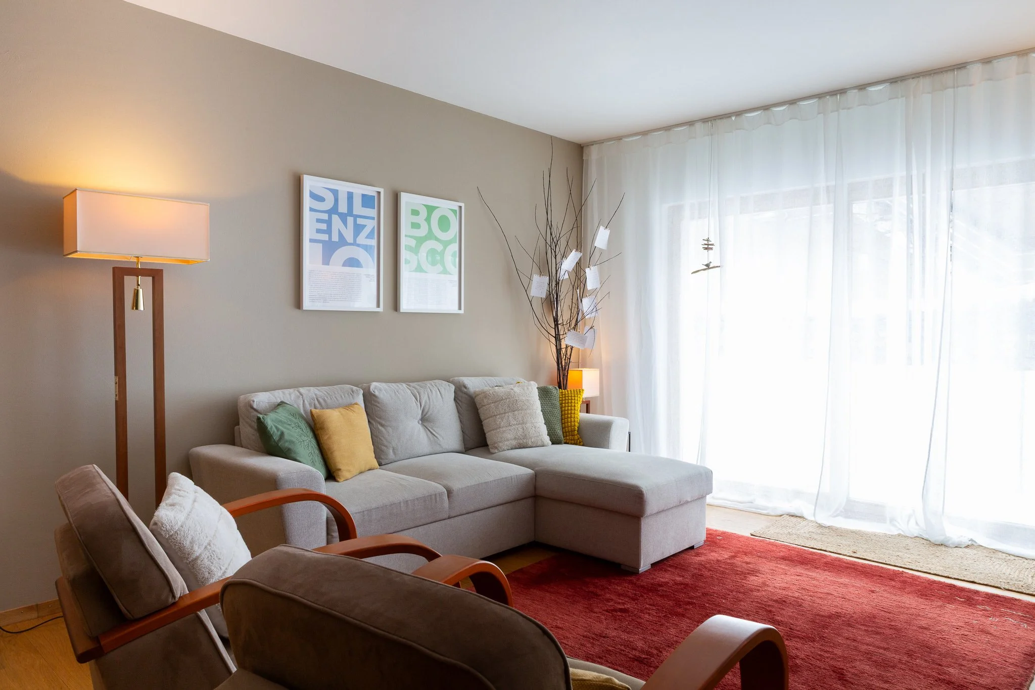 Living room with beige walls, white curtains, a beige sofa with colorful pillows, a wooden framed armchair, a red rug, a tall lamp, and wall art.