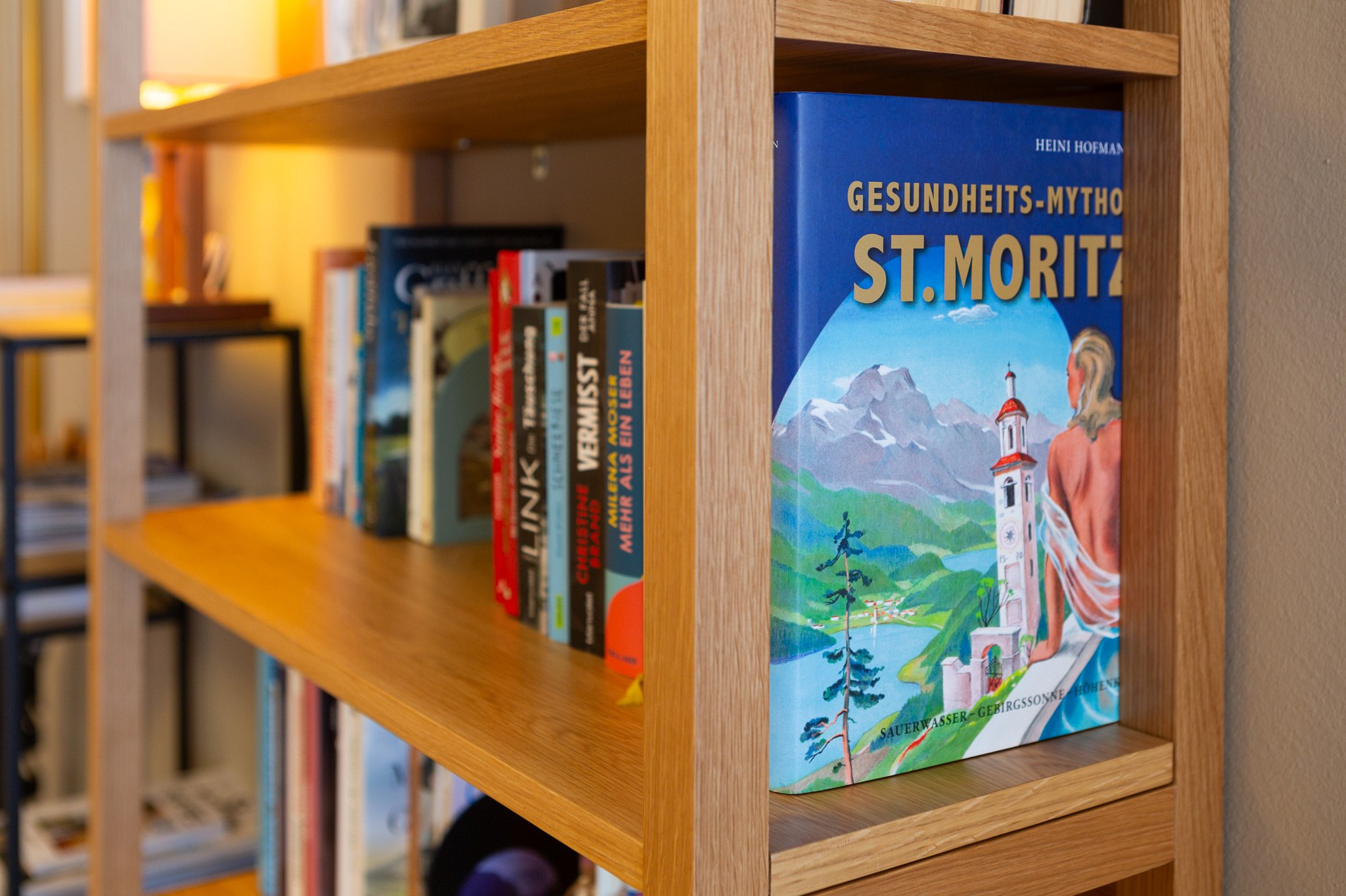 A wooden bookshelf with various books, prominently displaying a colorful book titled 'Gesundheits-Mythos St. Moritz' with illustrations of mountains, a lake, and a woman sitting on a bench.