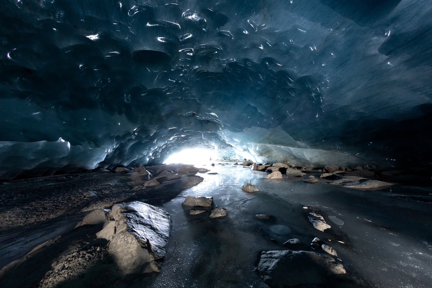 Ice cave with blue ice walls and rocks on the icy floor, light shining from the entrance in the distance.