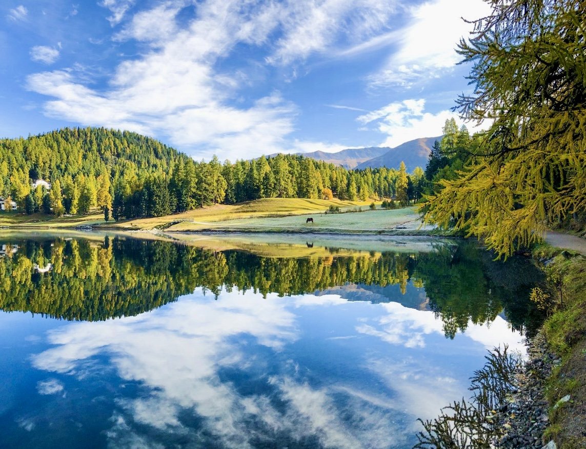 A serene lake reflects the blue sky with white clouds, surrounded by lush green trees and distant mountains.