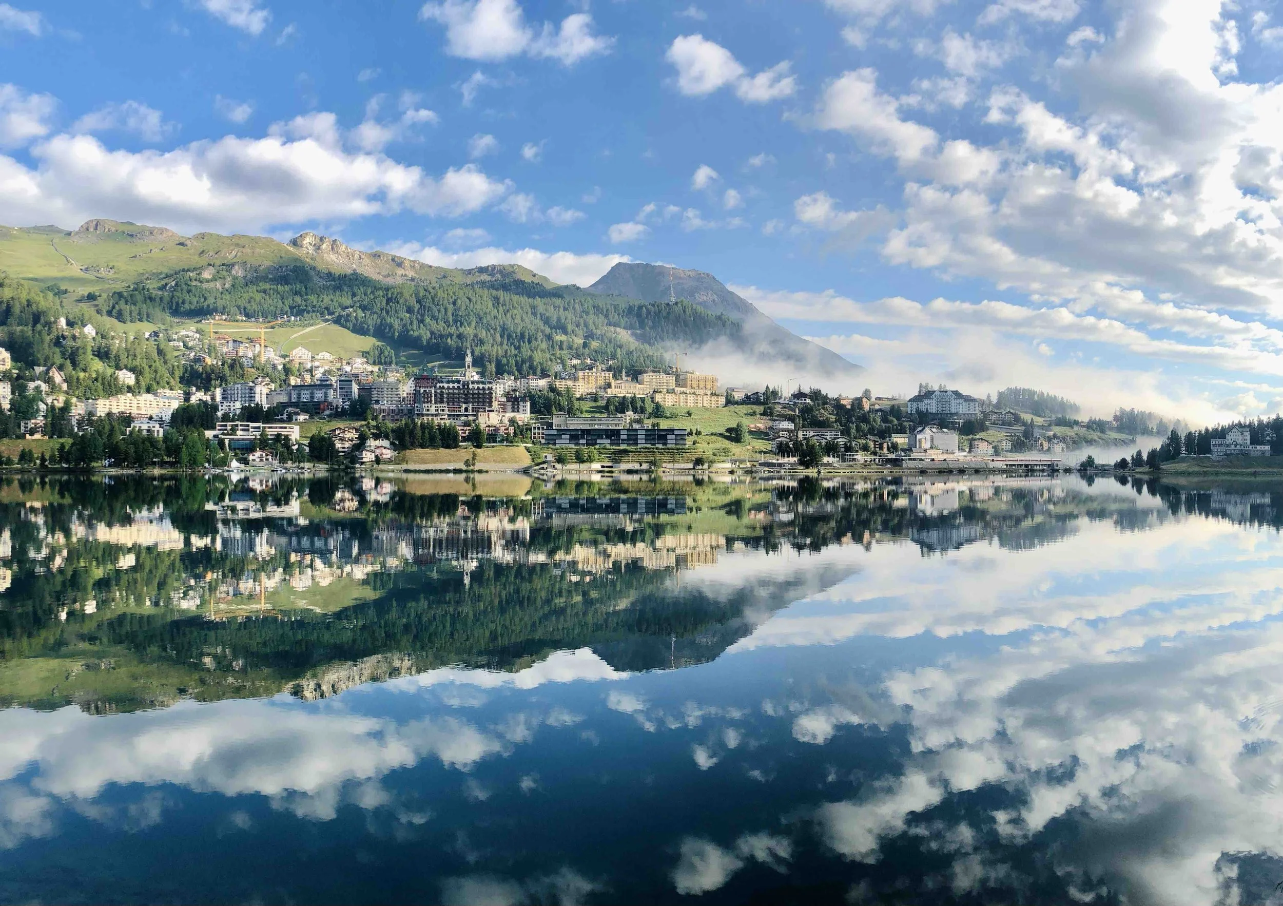 A scenic view of a town with multiple buildings and houses at the base of green mountain hills under a partly cloudy blue sky, with a reflection of the scene on a calm body of water.