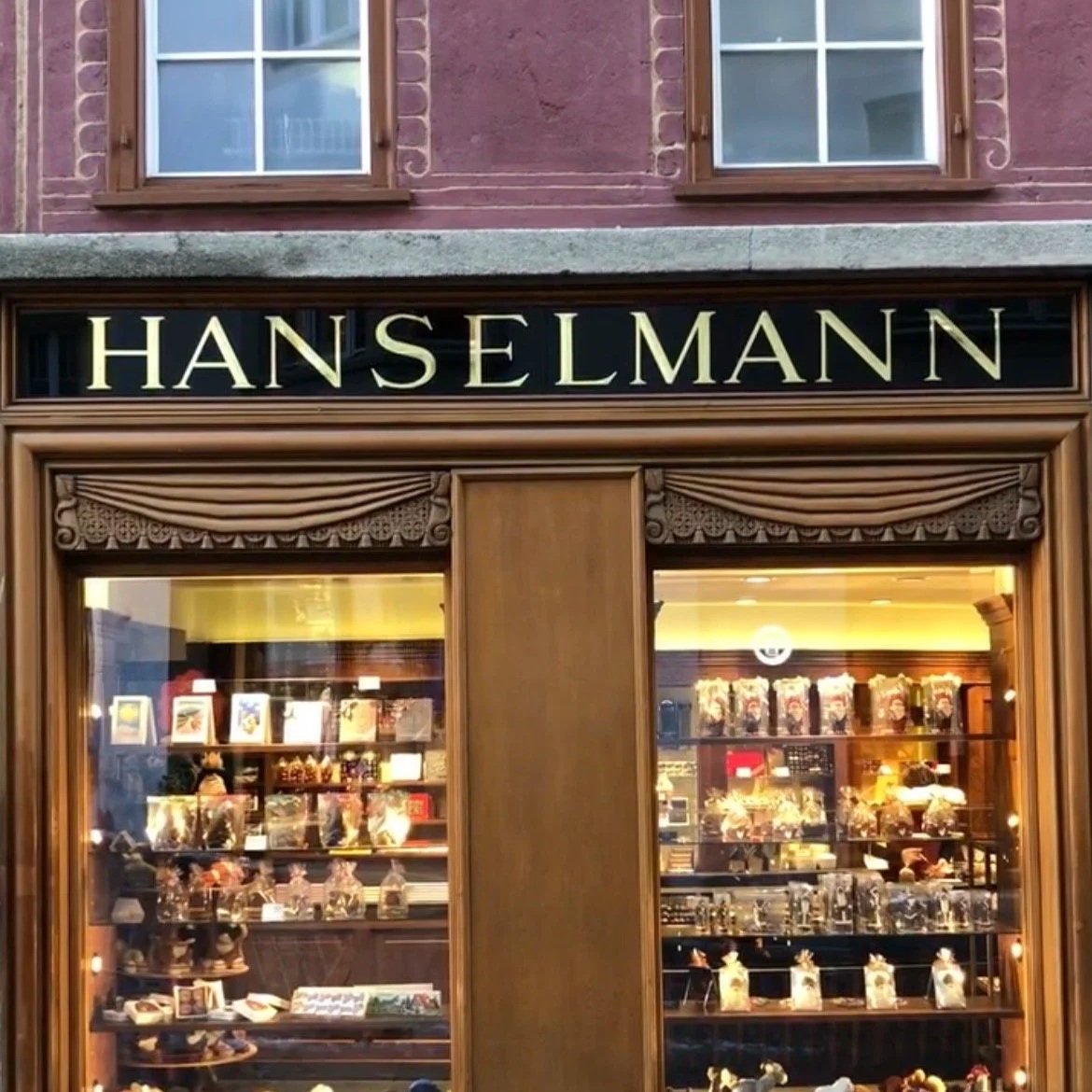  storefront of a shop named 'HanseIman' with wooden frame, glass display windows showcasing various products inside, above the shop is a building with two windows with wooden shutters.