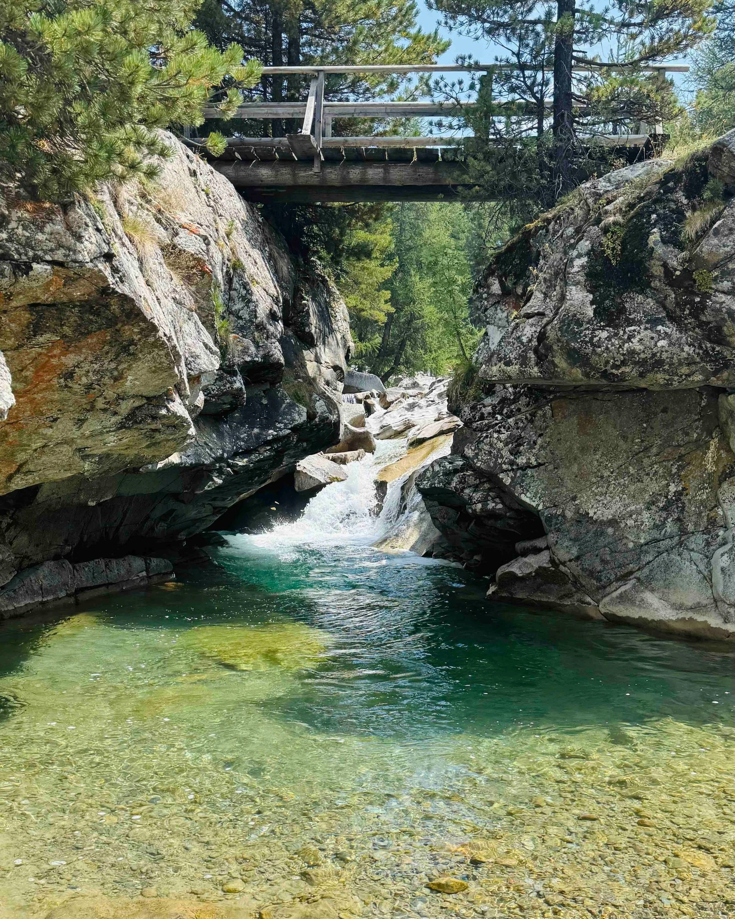 A rocky river with clear water flowing through a forested area with a wooden bridge overhead.