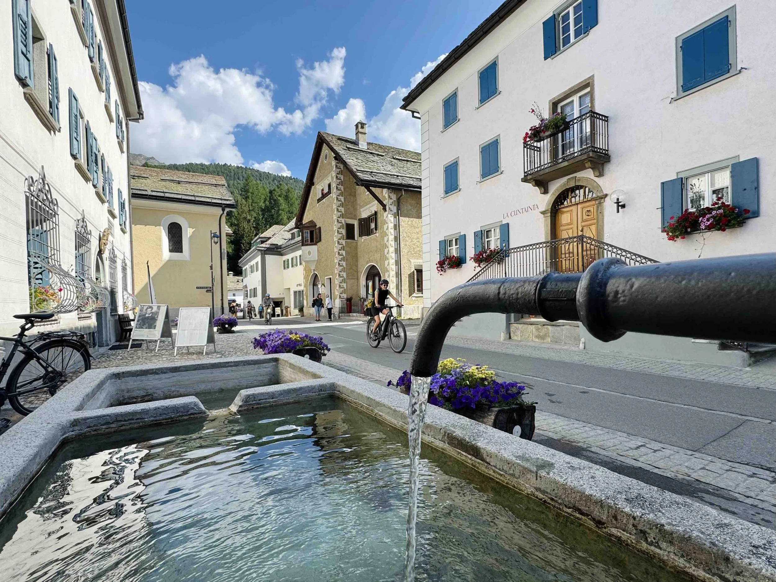 A street scene in a European town with historic buildings, people riding bikes, and a water fountain in the foreground under a blue sky with white clouds.