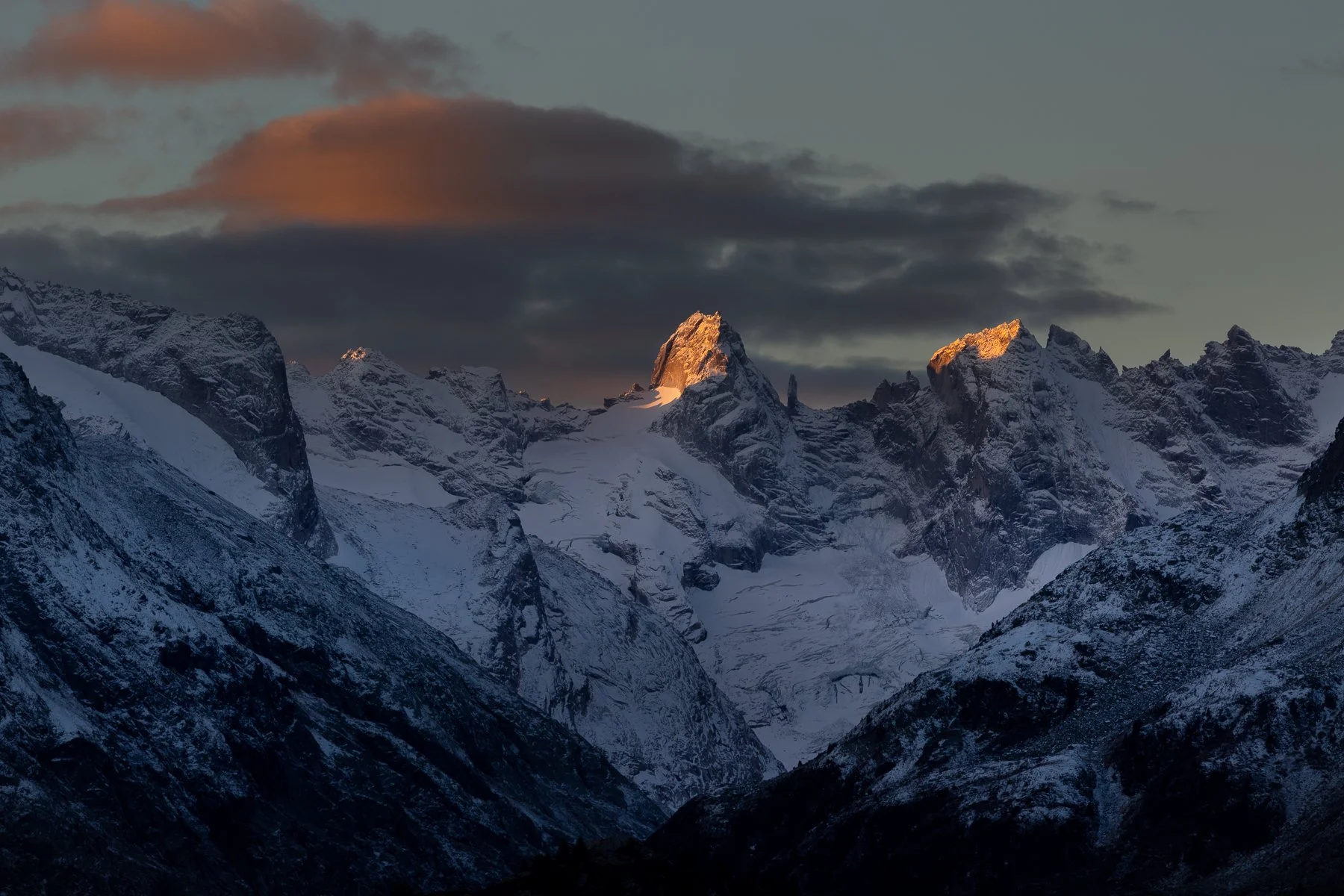 Snow-covered mountain peaks at sunset with dark clouds overhead.