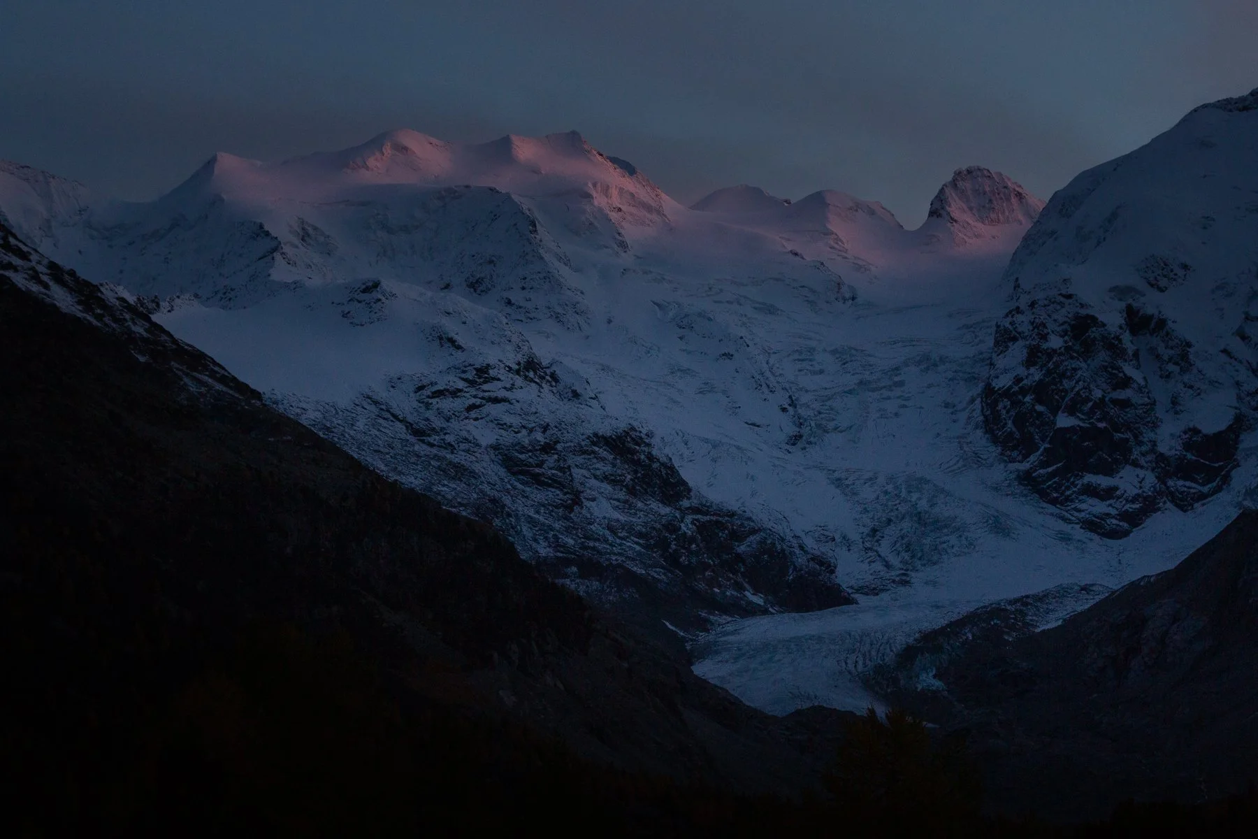 Snow-covered mountain range at dusk with a glacier in the valley.