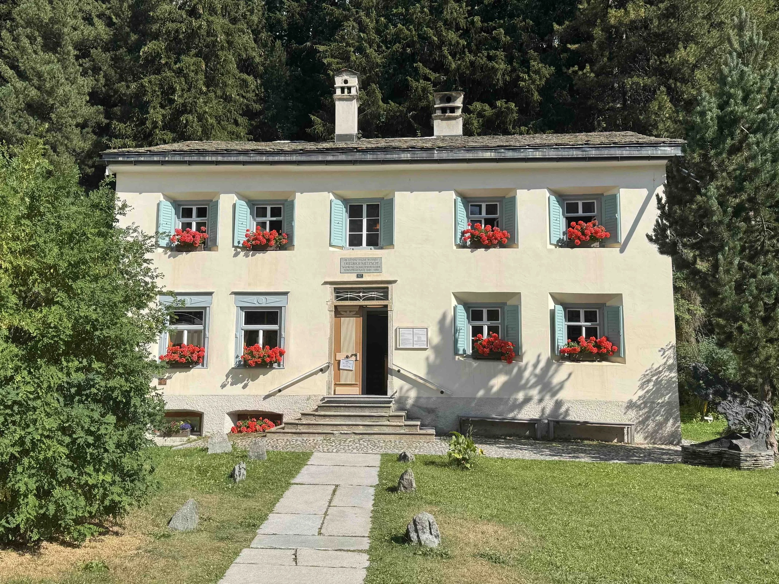 A two-story house with a light yellow facade, light blue shutters, and red flower boxes, surrounded by trees and a green lawn, with stone steps leading to the entrance.
