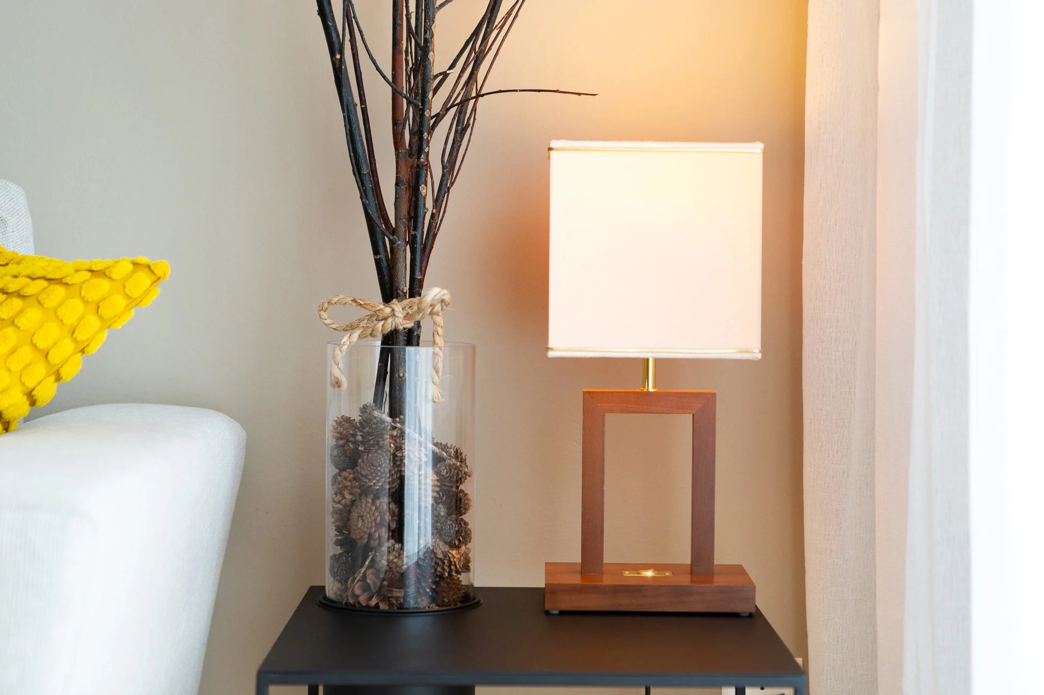 A table lamp with a rectangular wooden base and a beige fabric shade sits on a black table next to a tall glass vase filled with pinecones and branches, against a beige wall with a window and curtain to the right.