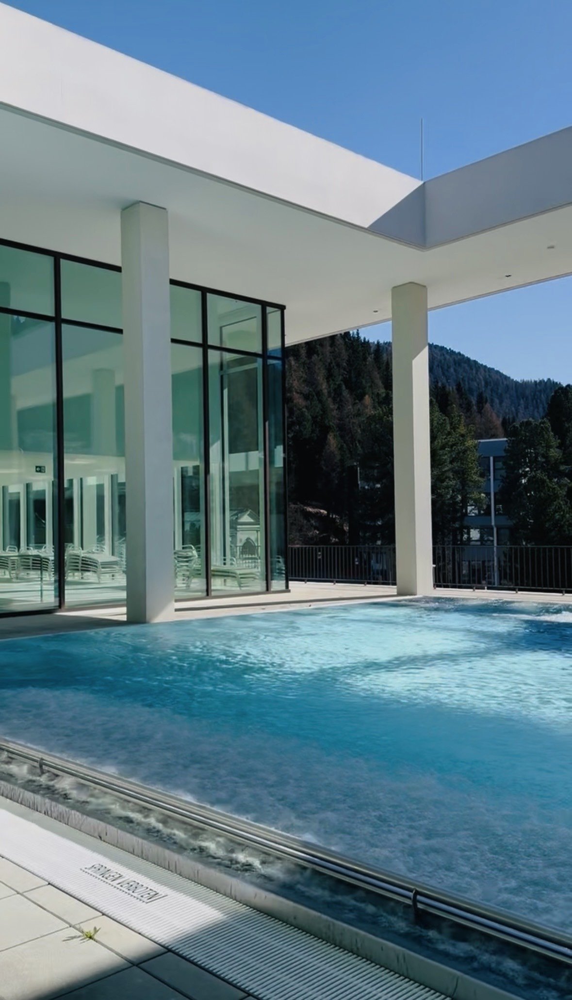 Modern outdoor pool area with glass walls, white columns, and mountain view in the background.