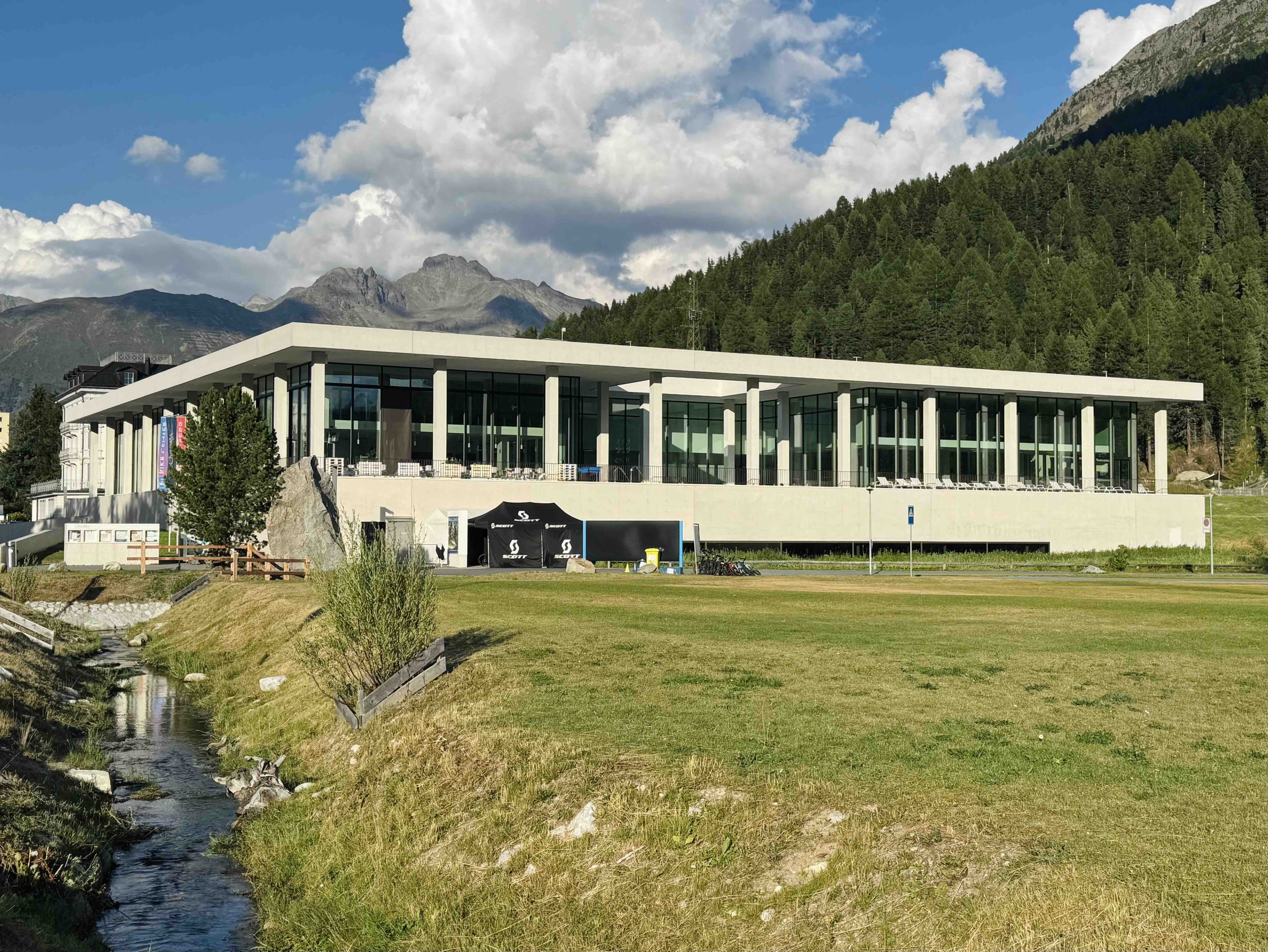 Modern building with large glass windows and flat roof, situated in a mountainous area with forested hills, a stream in the foreground, and mountains in the background.