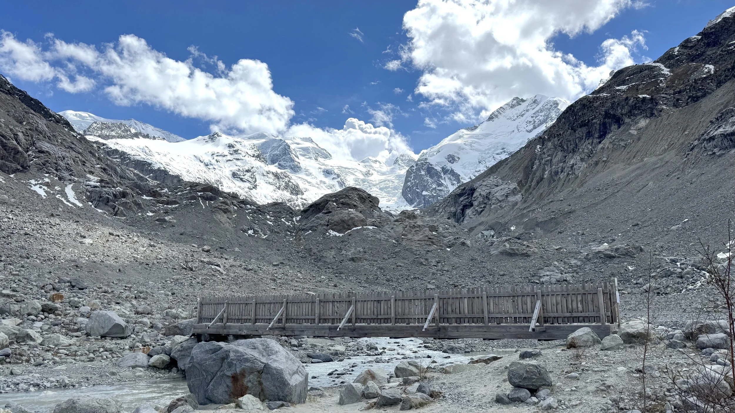 Snow-capped mountains with a partly cloudy blue sky, rocky terrain, and a wooden bridge over a small stream in a mountainous landscape.