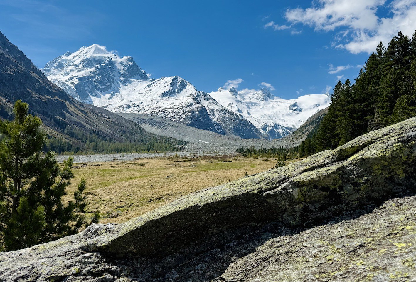 Snow-capped mountains in the distance with a green valley in the foreground, trees on the right side, and a large moss-covered rock in the lower right corner under a partly cloudy blue sky.
