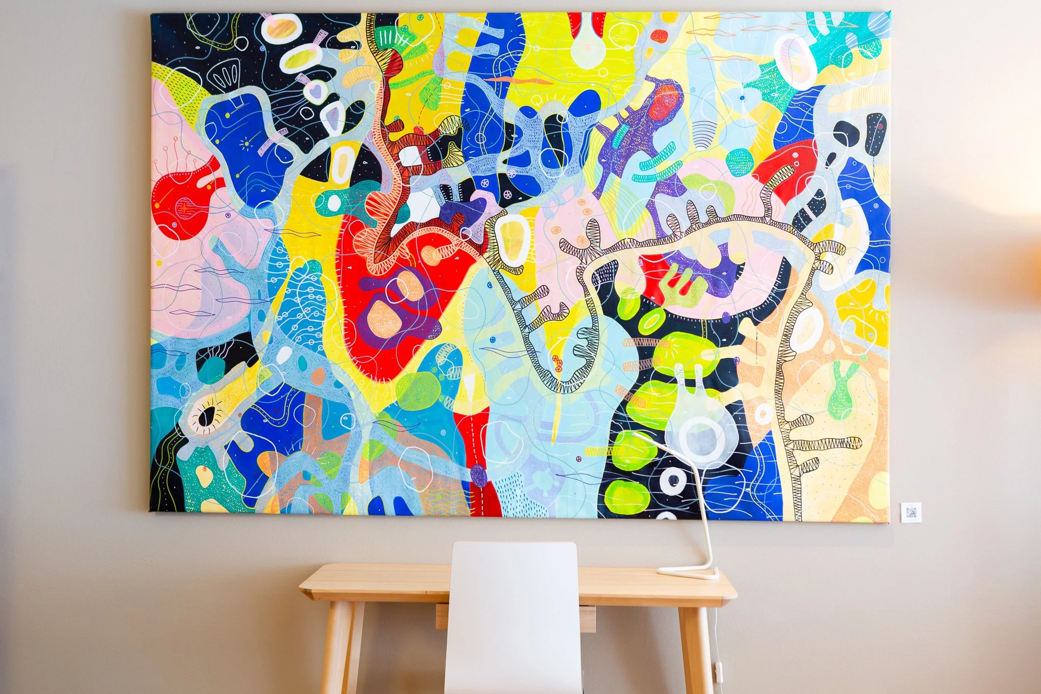 Colorful abstract modern wall art with various shapes and vibrant colors hung above a small wooden table with a white chair in front of it.