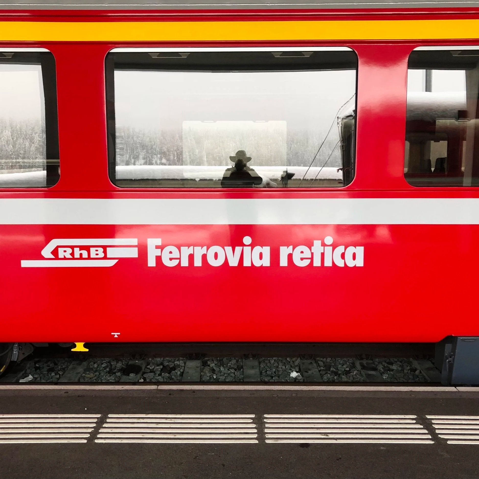 The image shows a red and white Railways hotel train car with windows. Through the window, a silhouette of a person wearing a hat can be seen, along with train equipment and snow-covered landscape outside.