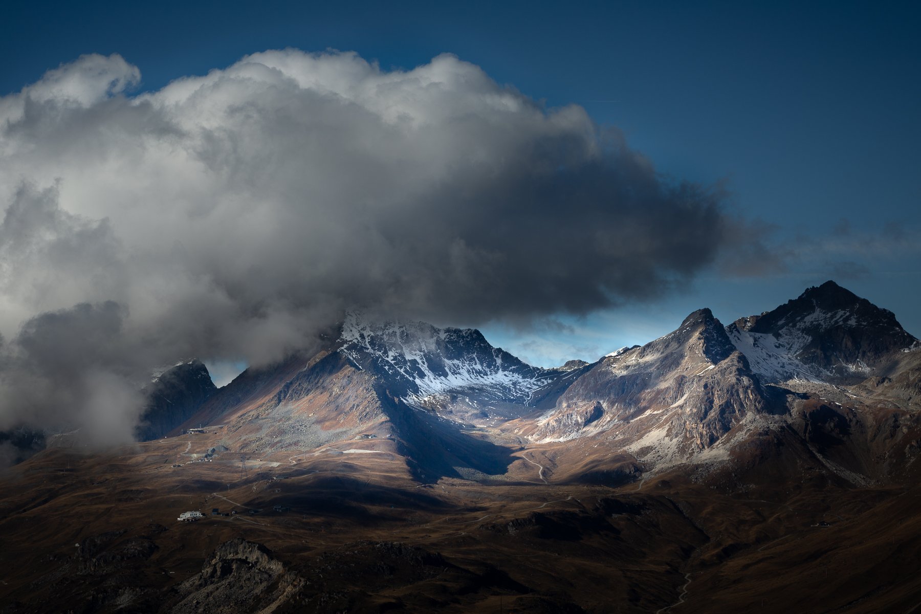 Mountain landscape with rugged peaks, some snow-capped, under a cloudy sky with patches of blue, and a few small buildings at the base.