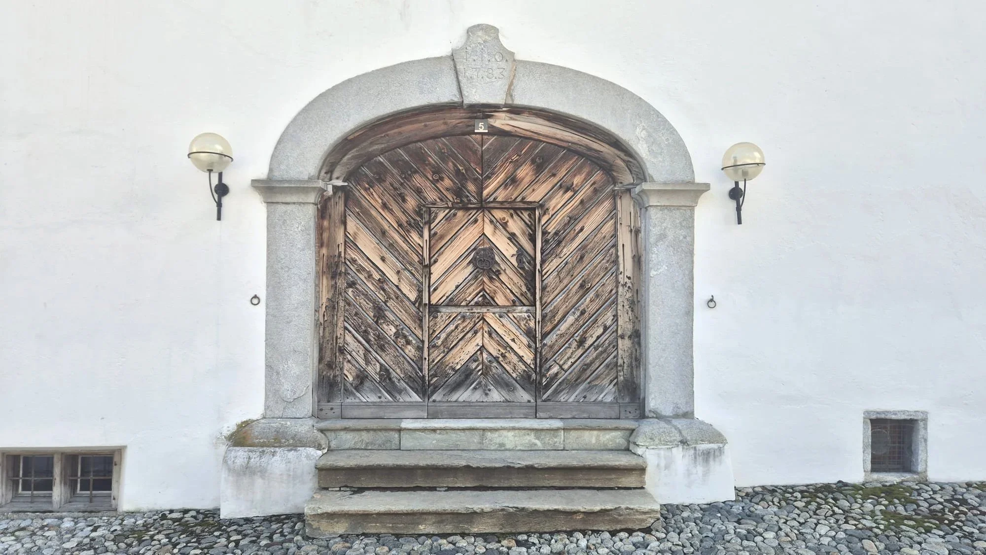 An arched wooden door with diagonal planks and ornate metal handles, framed by a stone archway, mounted on a white wall with two wall-mounted lamps on each side.