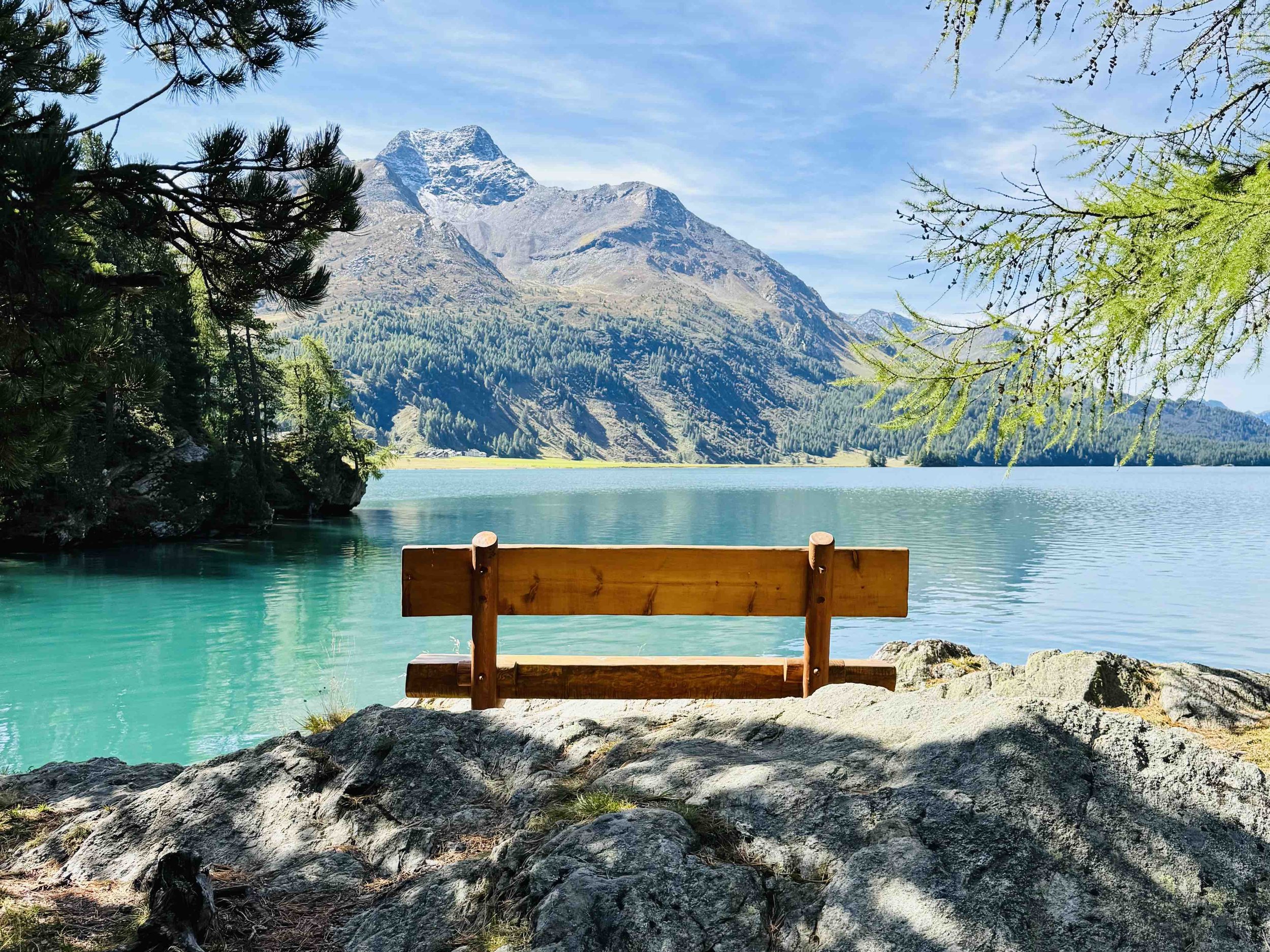 A wooden bench on rocks overlooking a turquoise lake, with mountains and trees in the background under a blue sky.