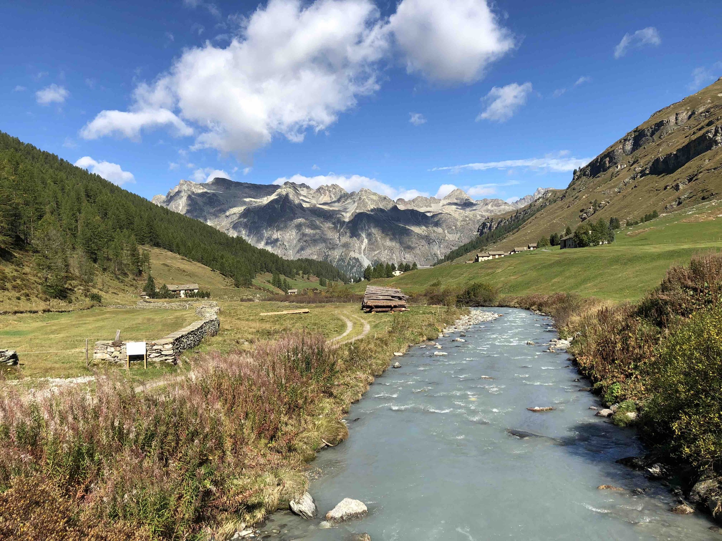 A scenic mountain landscape with a flowing river, green grassy fields, trees, small houses, and rugged mountain peaks under a blue sky with clouds.
