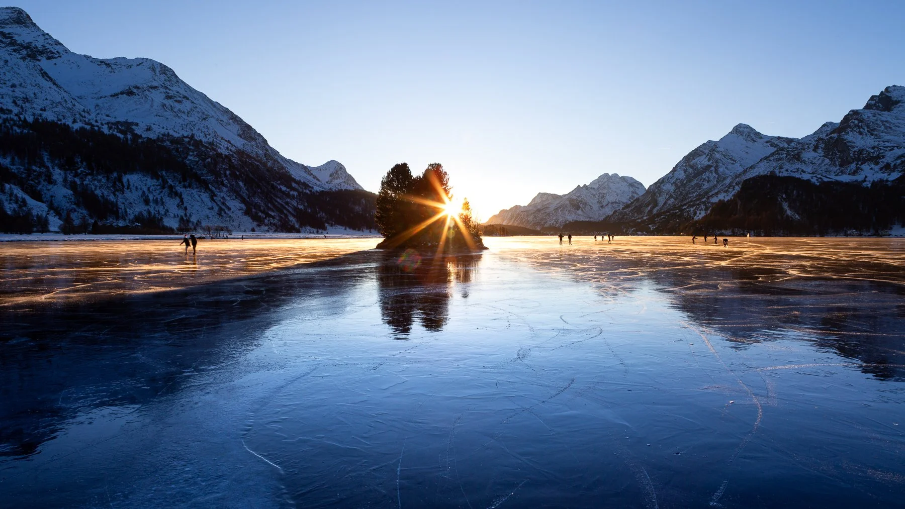 Sunset over a frozen lake surrounded by snow-covered mountains, with several people ice skating and a small island with trees in the center.