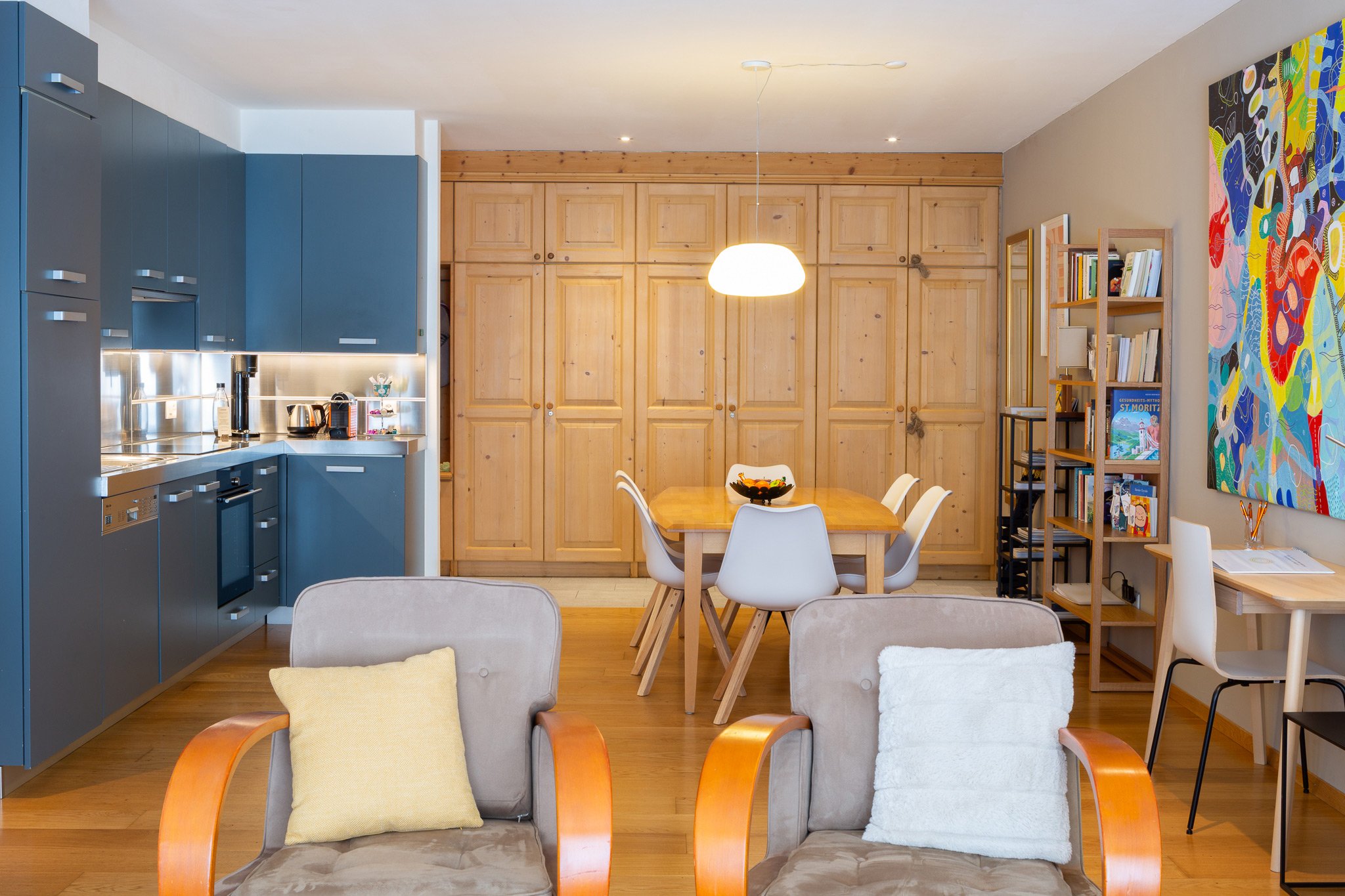 Living room with kitchen on the left, wooden dining table in the center with white chairs, blue and wooden cabinets, colorful large wall art, bookshelves, and beige armchairs with yellow and white pillows.