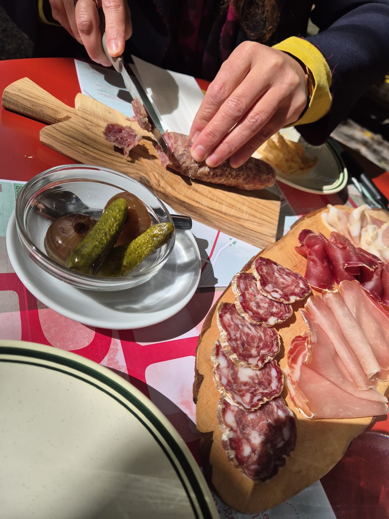 Close-up of a person slicing cured meats on a wooden board, with a bowl of pickles and an assortment of cured meats on a wooden serving board nearby, on a red tablecloth with menus underneath.