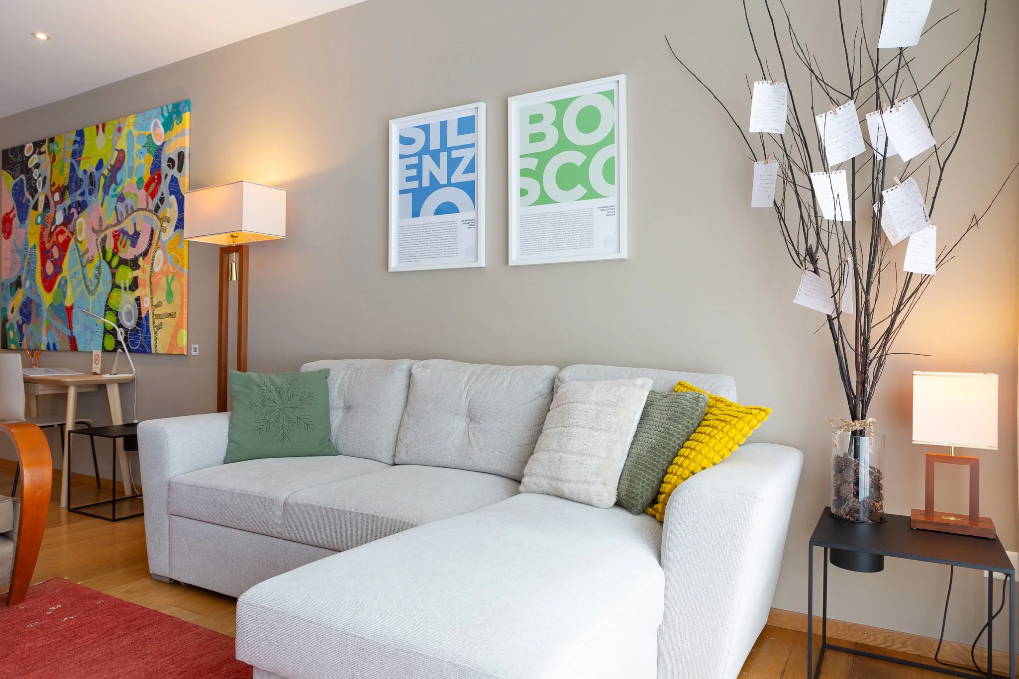 Modern living room with beige wall, white sofa with green, beige, and yellow pillows, colorful abstract painting, framed blue and green posters, floor lamp, and decorative tree with notes hanging from branches.