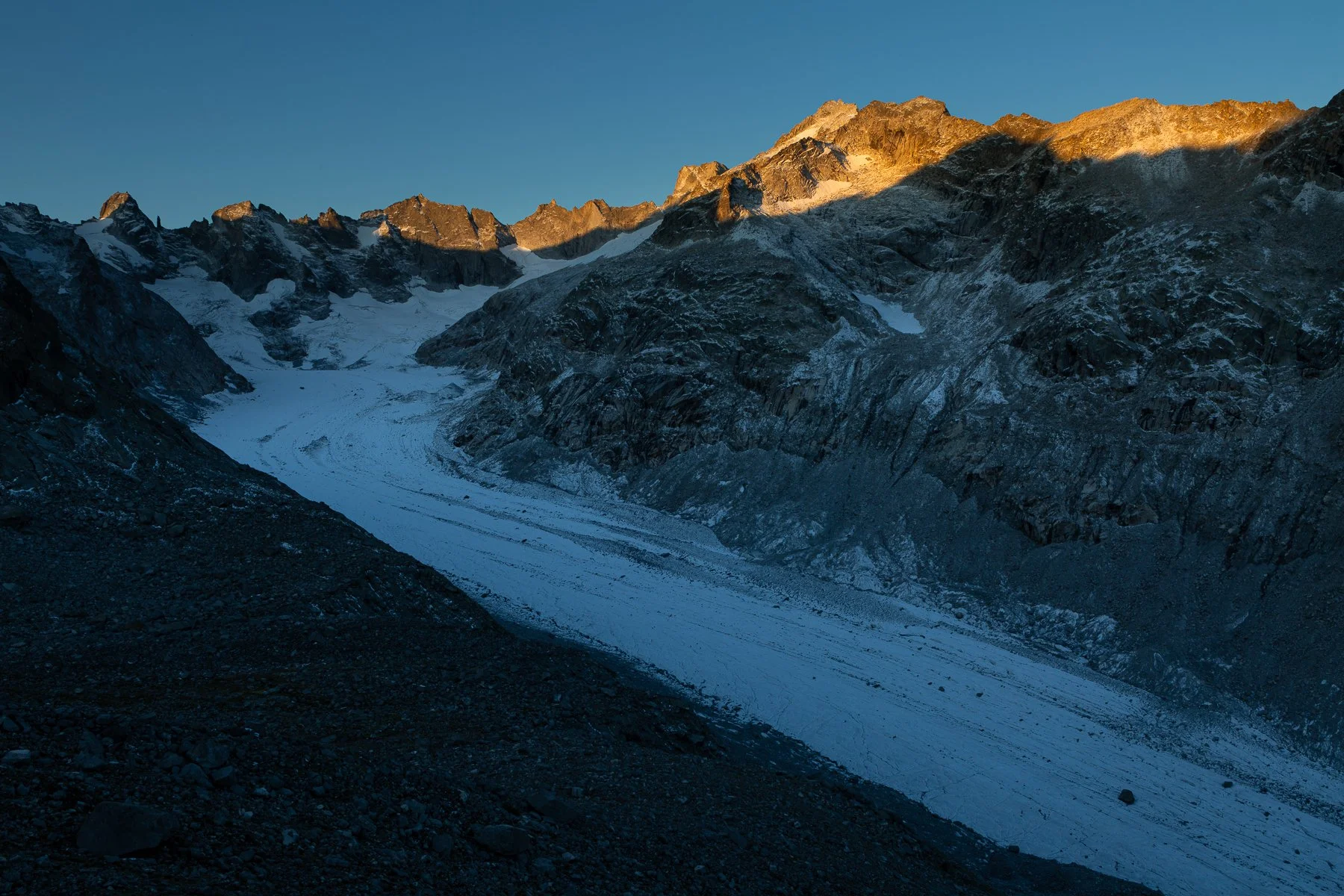 Snow-covered glacier descending between rugged mountain peaks under a clear blue sky, with sunlight illuminating the mountain summits.
