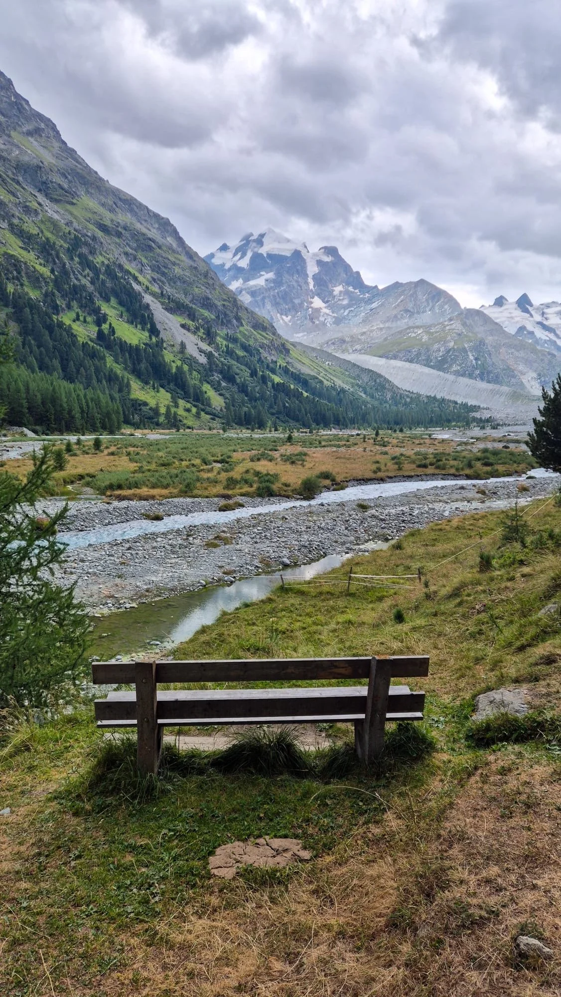 View of a mountain valley with green slopes, a river flowing through the valley, and snow-capped mountains in the background. A wooden bench sits in the foreground.