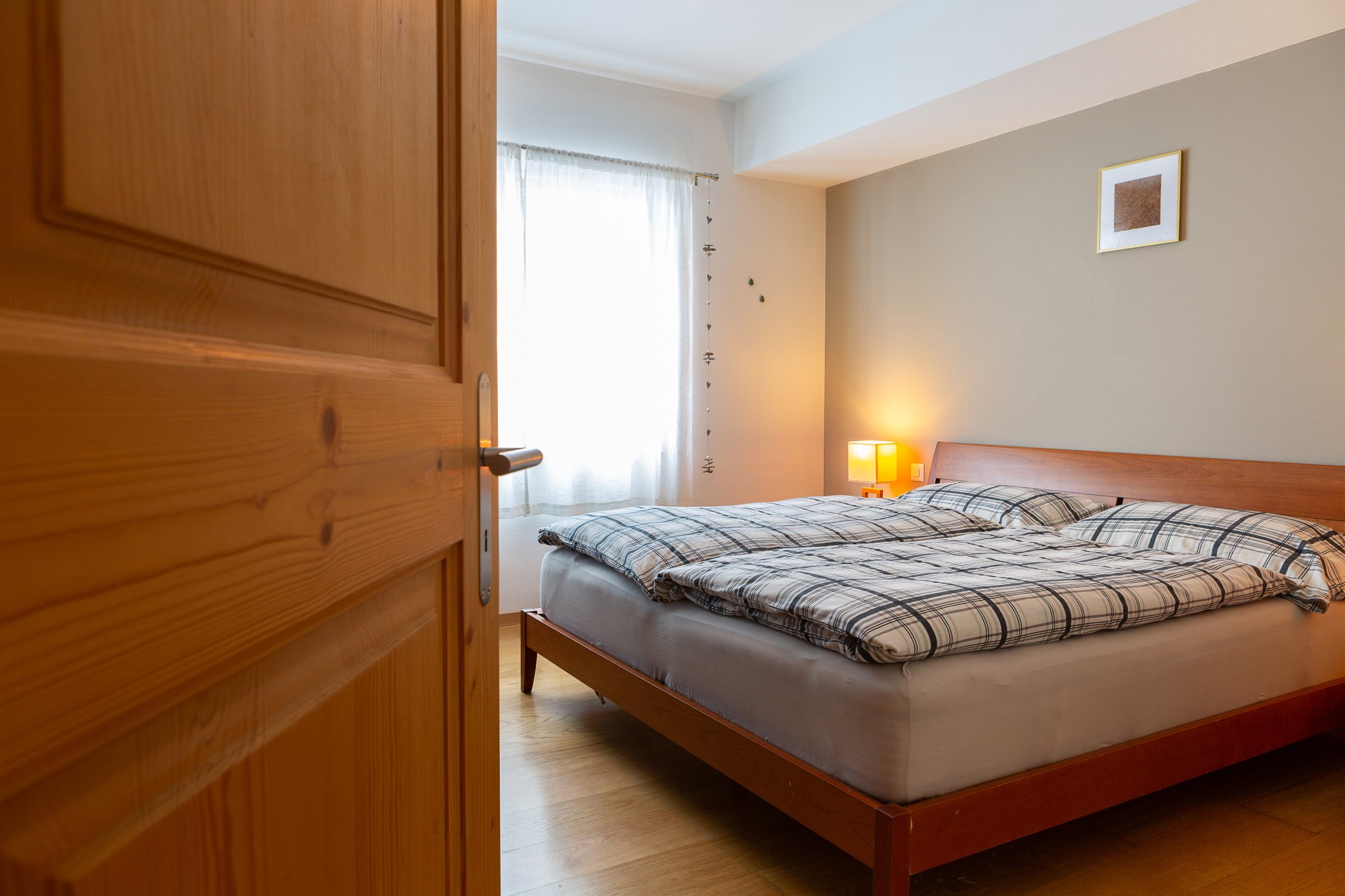 A bedroom with a wooden bed, plaid bedding, a bedside table with a yellow lamp, framed artwork on the wall, a window with sheer curtains, and a wooden door partially open.