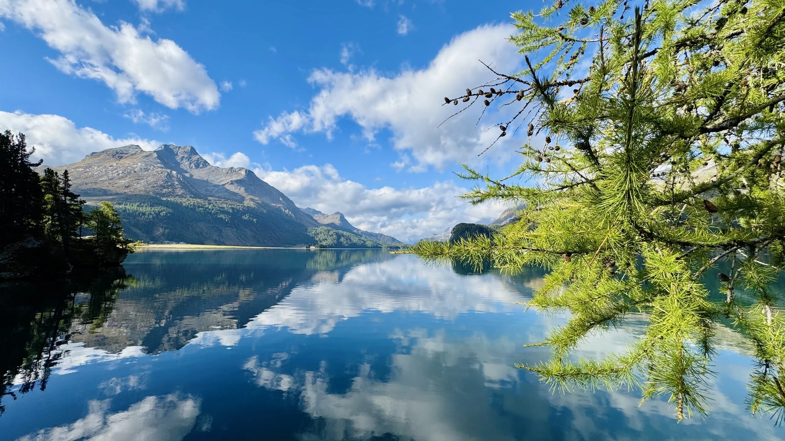 A serene mountain lake scene with a clear blue sky, white clouds, tall mountains in the background, and green trees and foliage reflecting on the calm water.