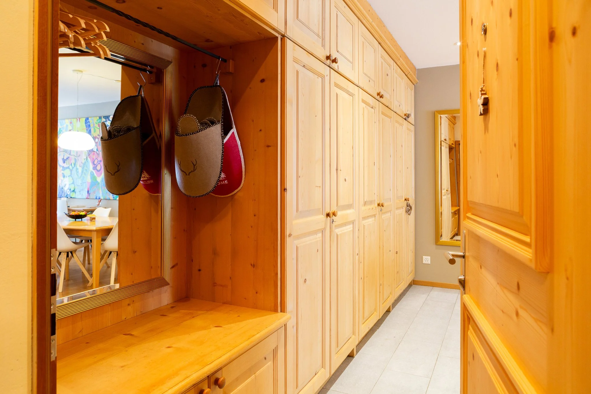 Wooden hallway with storage cabinets and hanging slippers, mirror reflecting dining area with colorful art.