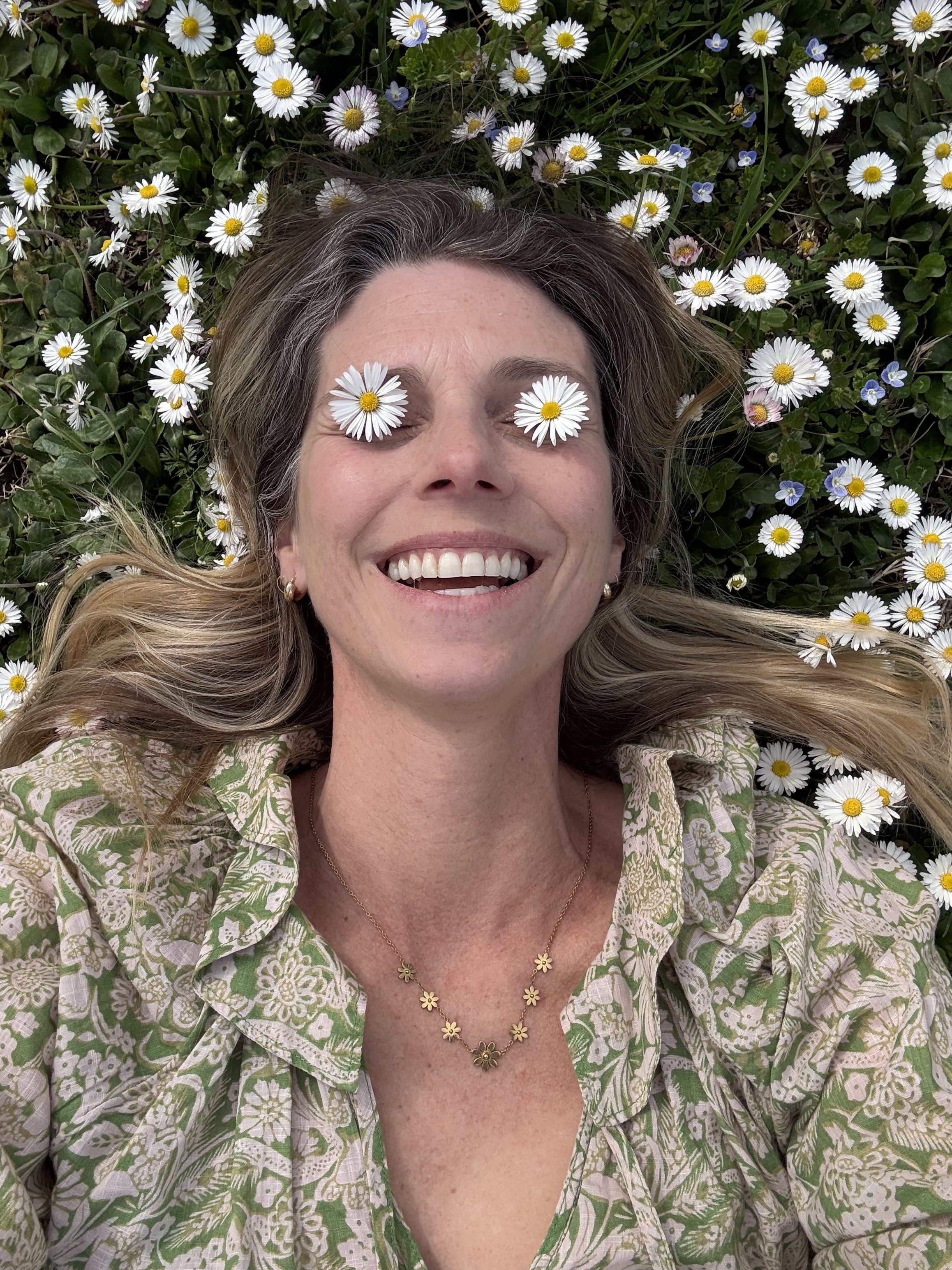 A woman lying on the grass surrounded by white daisies and small blue flowers, smiling with flowers over her eyes.