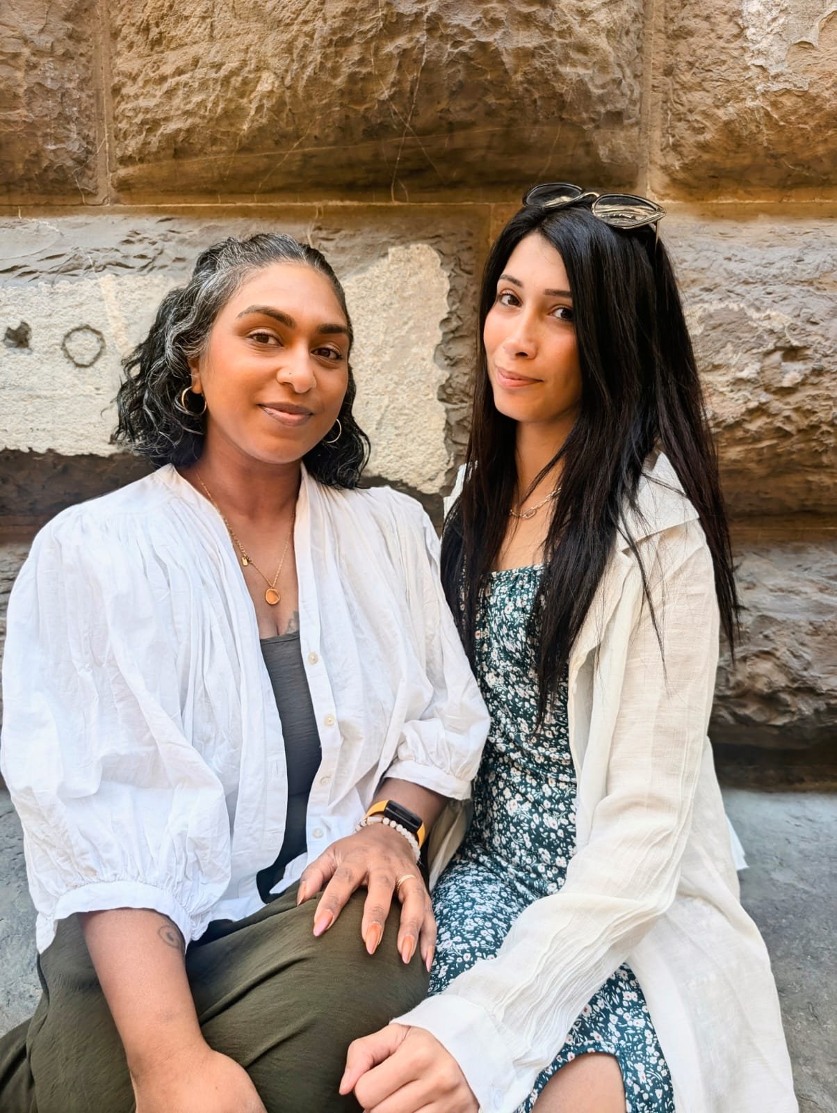 Two women sitting close together against a stone wall, smiling at the camera. The woman on the left has shoulder-length dark curly hair, wearing a white button-up shirt and gold jewelry. The woman on the right has long straight dark hair, wearing a cream-colored jacket over a patterned dress, with sunglasses on her head.