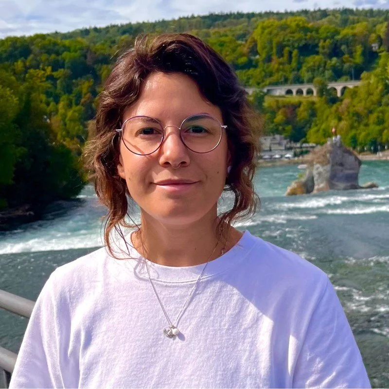 A young woman with short wavy hair and glasses smiling outdoors near a river, with lush green trees and a bridge in the background.