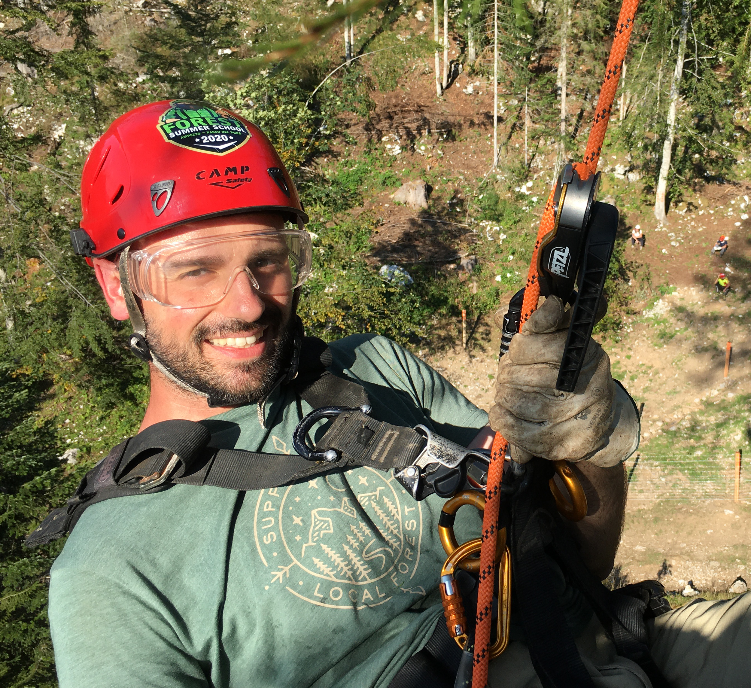 A man wearing a red climbing helmet and safety goggles, holding an orange climbing rope with a black and silver safety mechanism, smiling outdoors in a forested area.