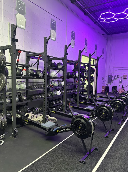 Row of rowing machines in a gym with weight racks and medicine balls, illuminated by purple LED lighting.