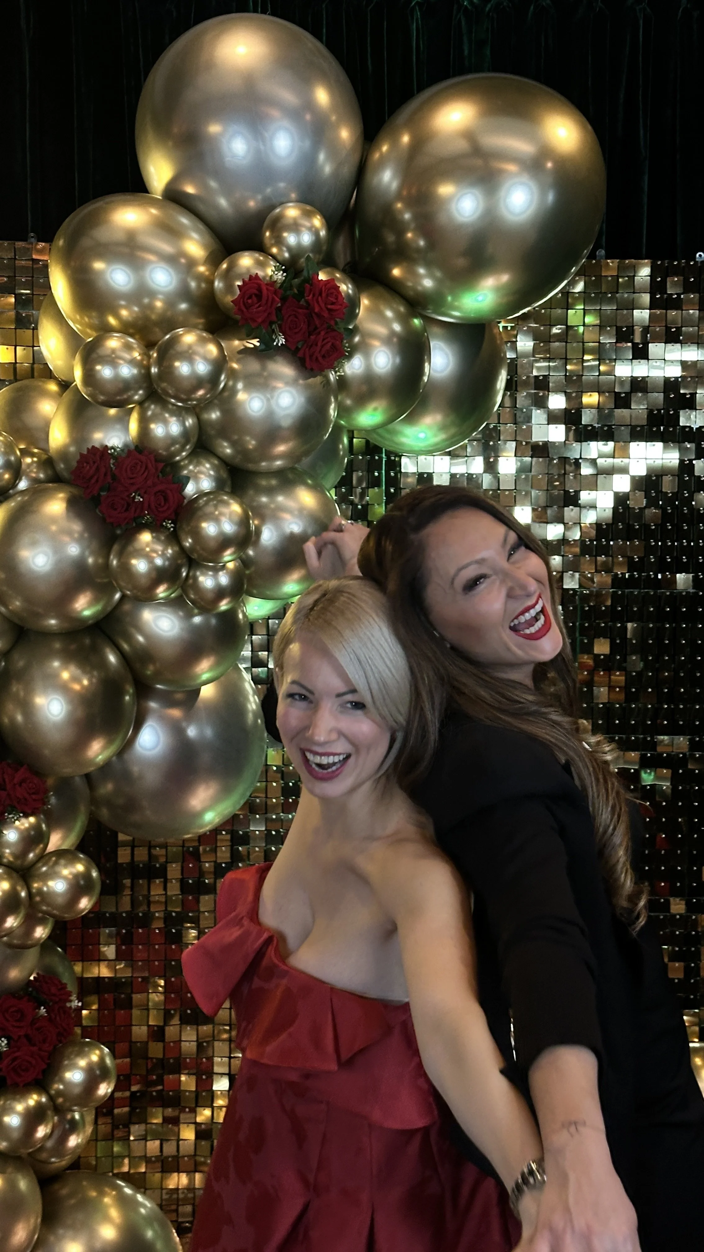 Zwei lachende Frauen vor einer Dekoration mit goldenen Luftballons und roten Rosen.