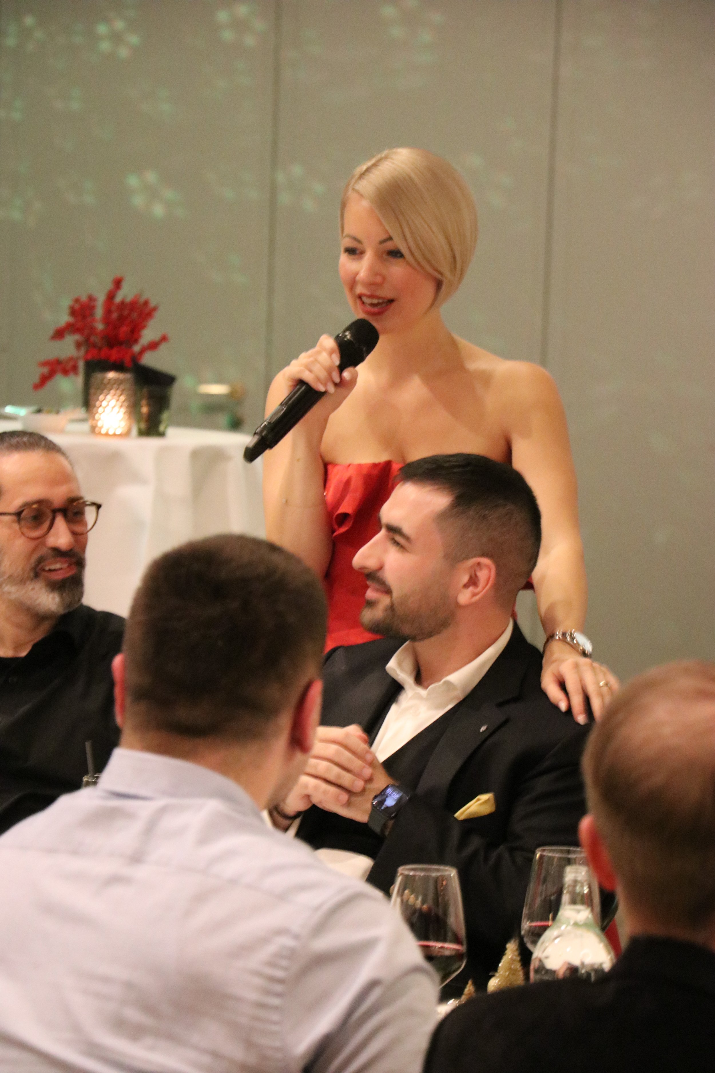 Eine junge Frau mit kurzen blonden Haaren, die in einem roten Kleid mit einem Mikrofon bei einer Feier spricht. Um sie herum sitzen Männer, die zuhören und lachen, bei einem festlichen Abendessen mit Wein. In der Hintergrunddecke gibt es Lichtreflexe