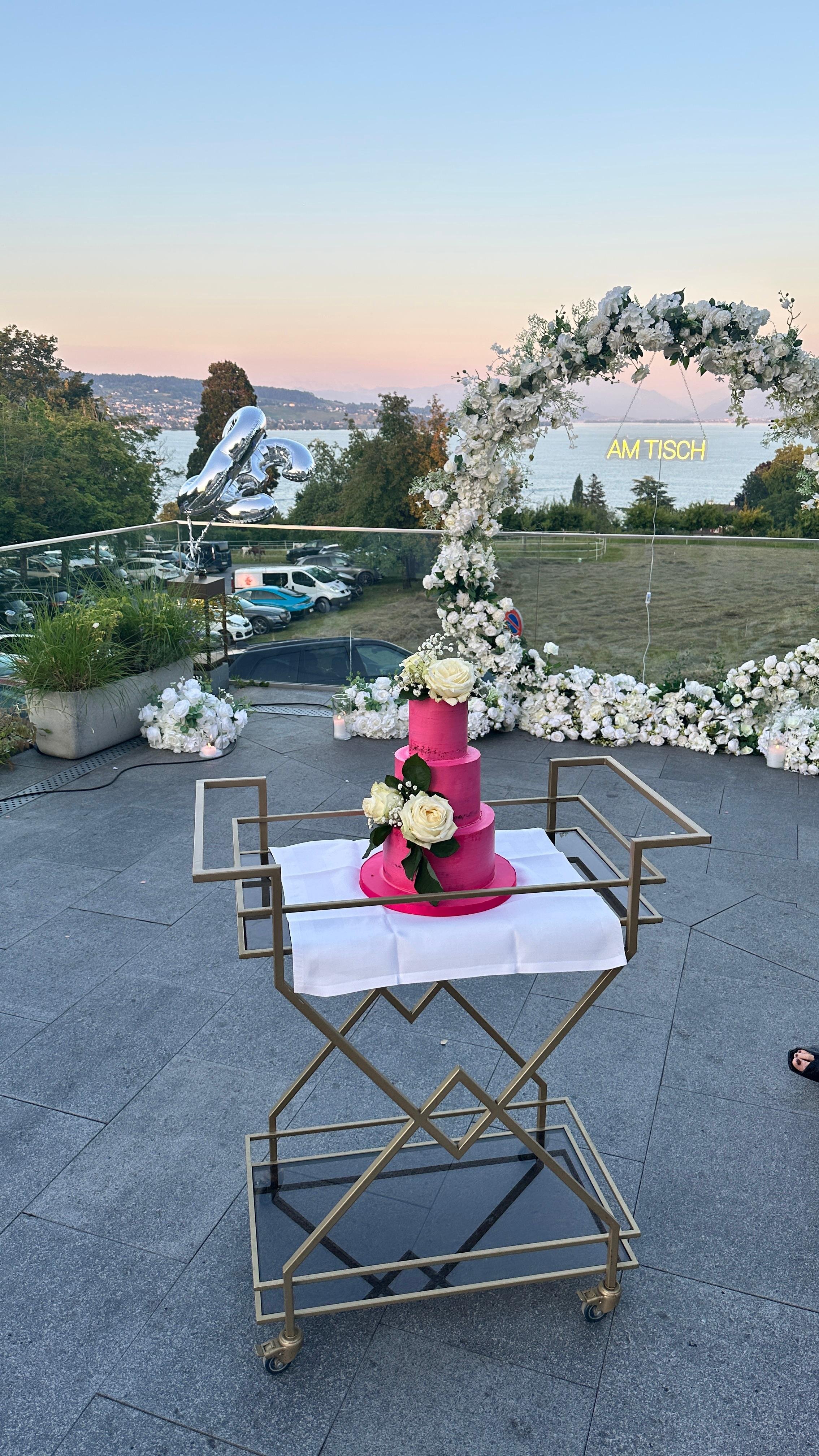 Ein dreistöckiger pinker Hochzeitstorte mit weißen Blumen, auf einem beweglichen Tablett, im Freien bei Sonnenuntergang mit Dekoration aus weißen Blumen und einem Wasserblick im Hintergrund.