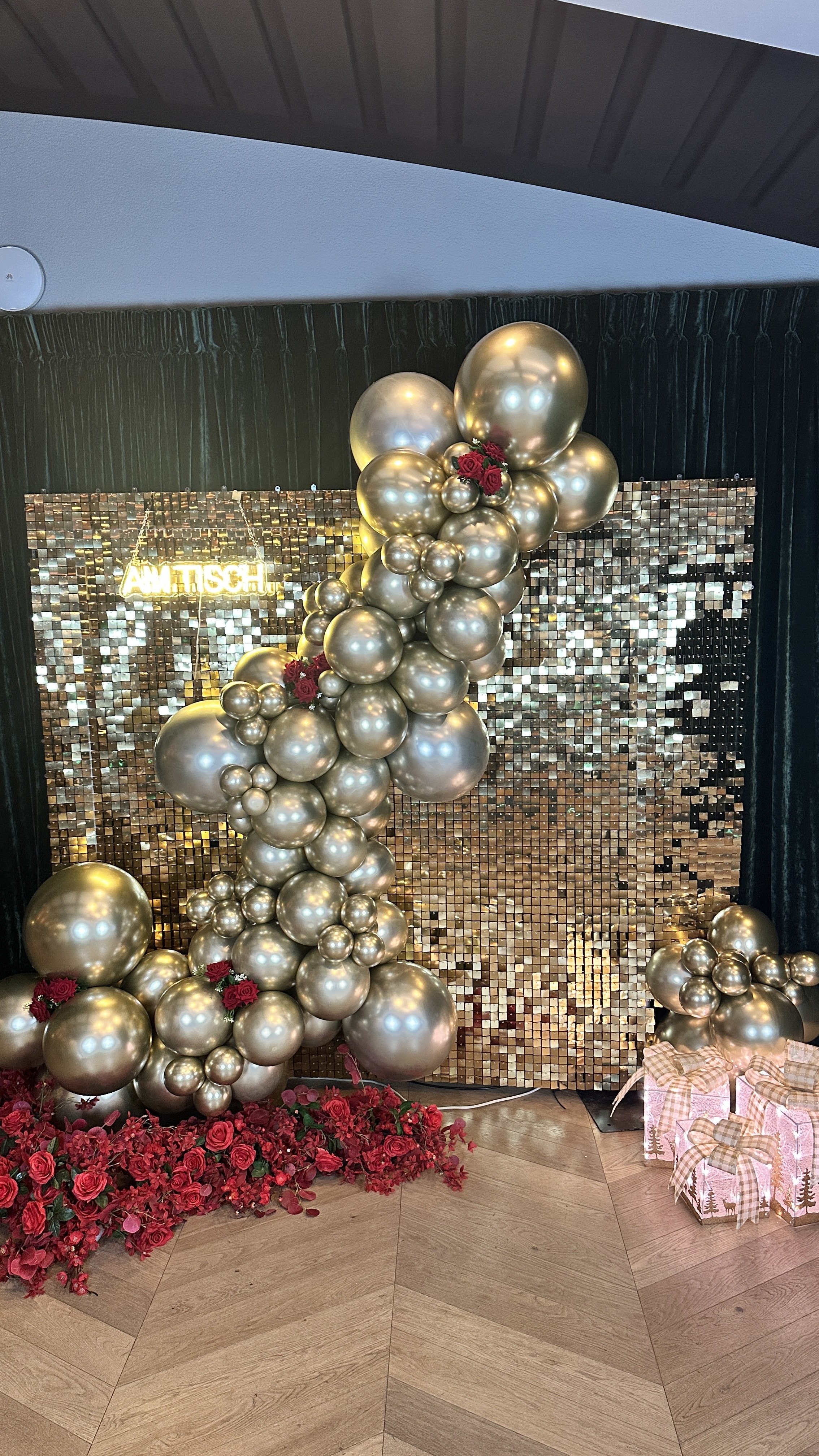 Dekoration mit goldenen Ballons, roten Rosen, roter Blumen und geschenkverpackungen vor einer goldenen Mosaikwand, mit einem leuchtenden Schild, das 'Amitisch' sagt.