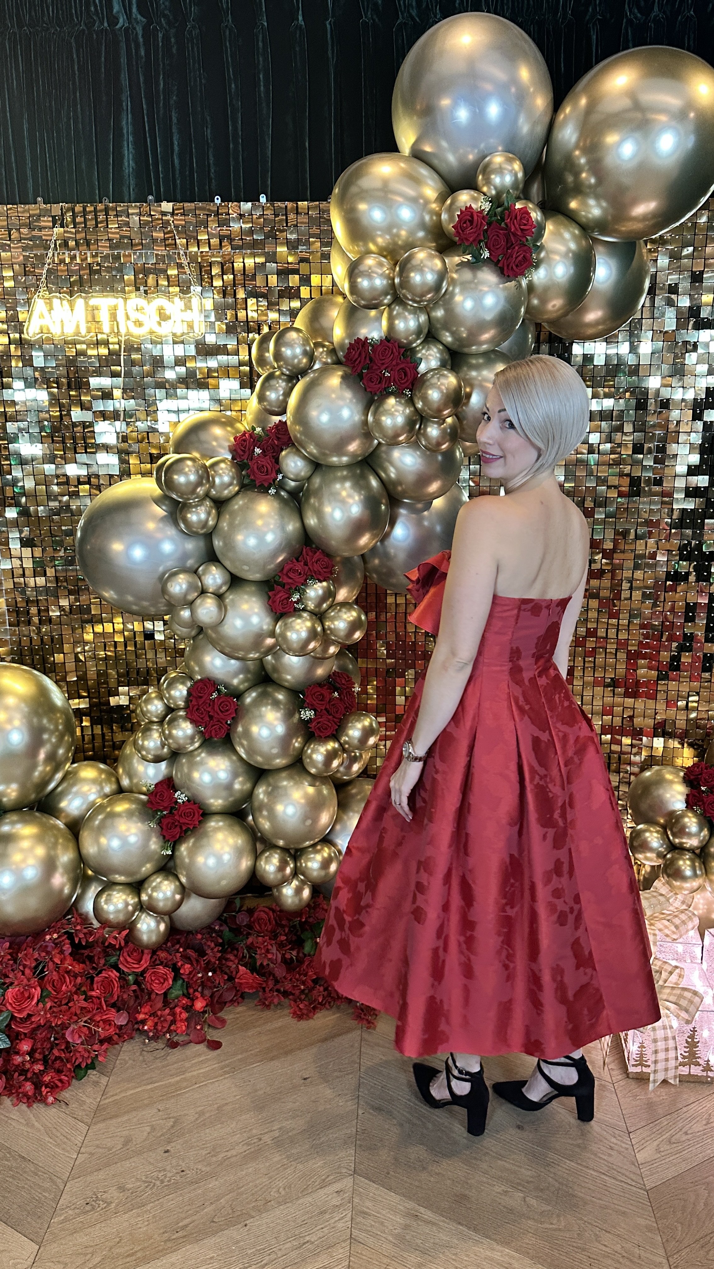 Eine Frau in einem roten, eleganten Kleid mit hohen Schuhen steht vor einer festlichen Dekoration aus goldenen Ballons, roten Rosen und einer glitzernden Wand. Im Hintergrund leuchtet eine neonartige Plakette.