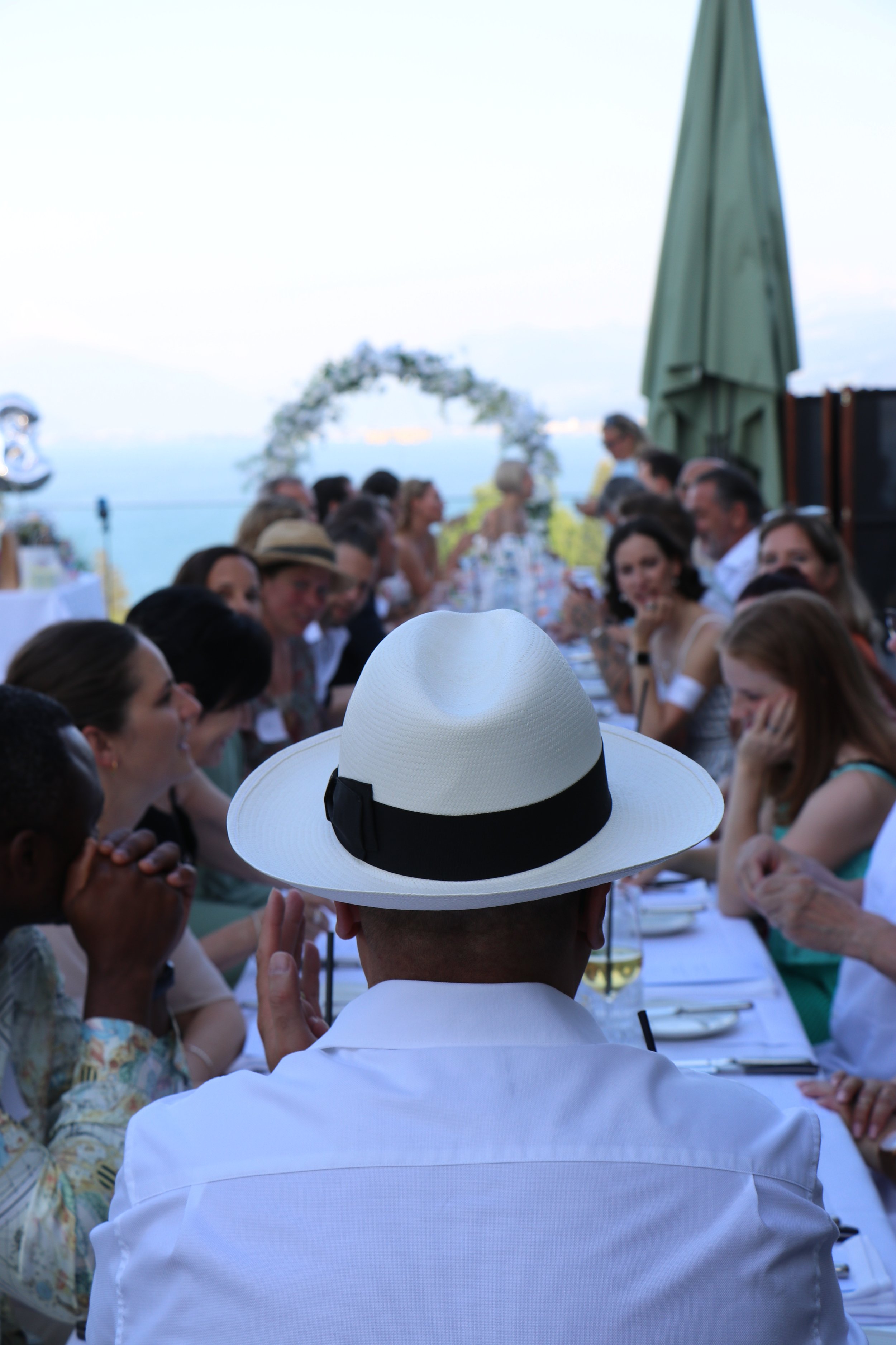 Menschen sitzen an einem langen Tisch im Freien bei einer Erfrischung oder einem Essen, im Hintergrund ist ein blauer Himmel und Wasser zu sehen, viele sind in sommerlicher Kleidung.
