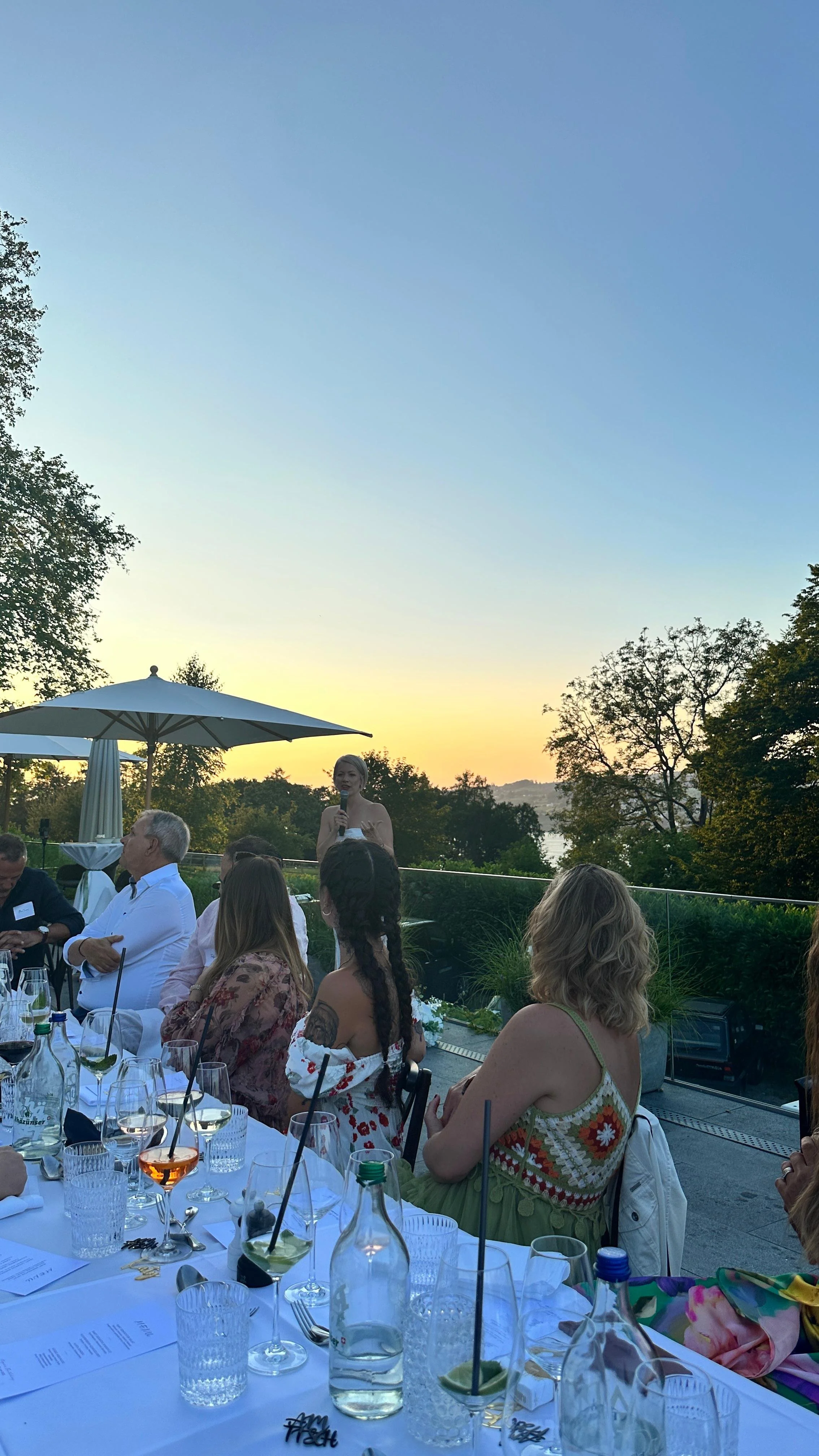 Menschen sitzen bei einem Abendessen im Freien, während eine Frau mit einem Mikrofon spricht, im Hintergrund ein Sonnenuntergang und Bäume.