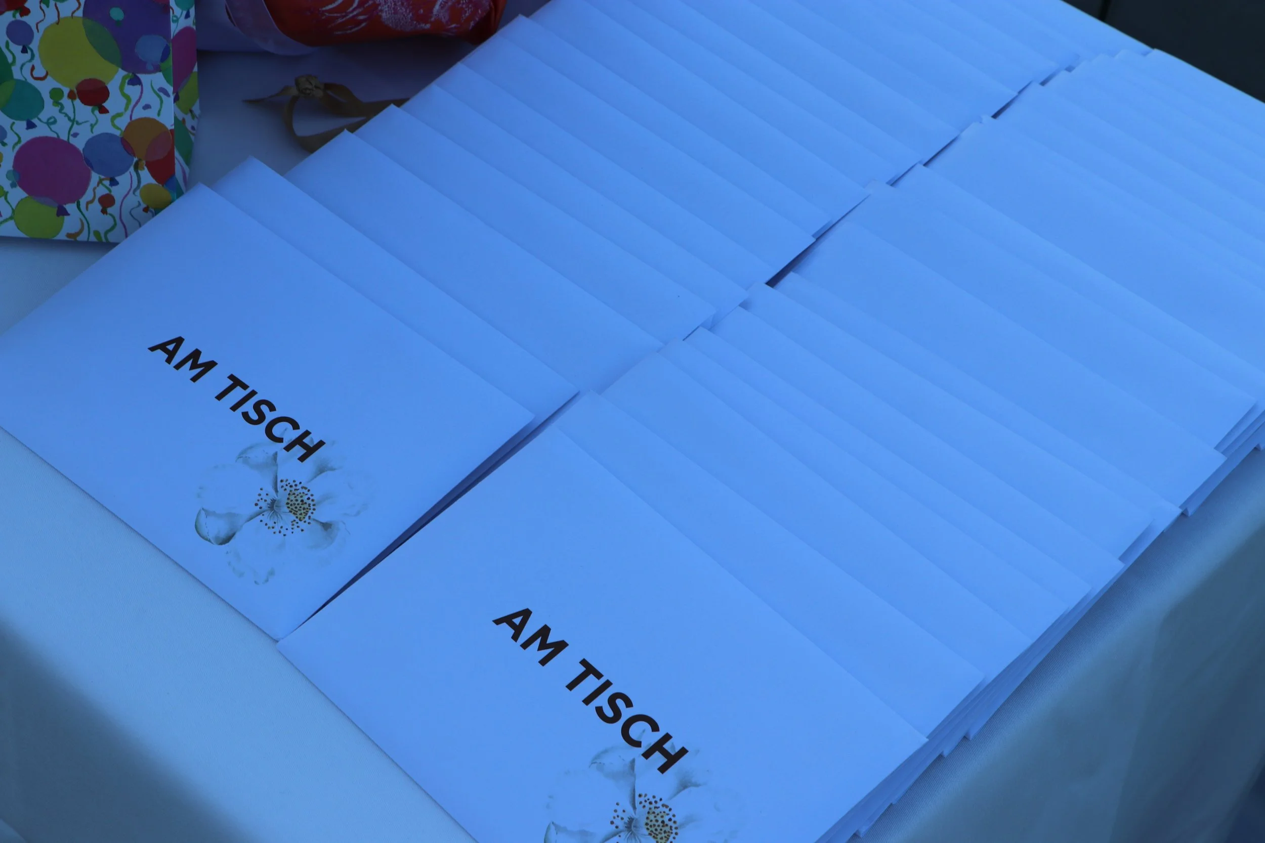 Mehrere weiße Karten mit dem Aufdruck "AM TISCH" liegen auf einem Tisch. Im Hintergrund sind partytaugliche Dekorationen und Süßigkeiten sichtbar.