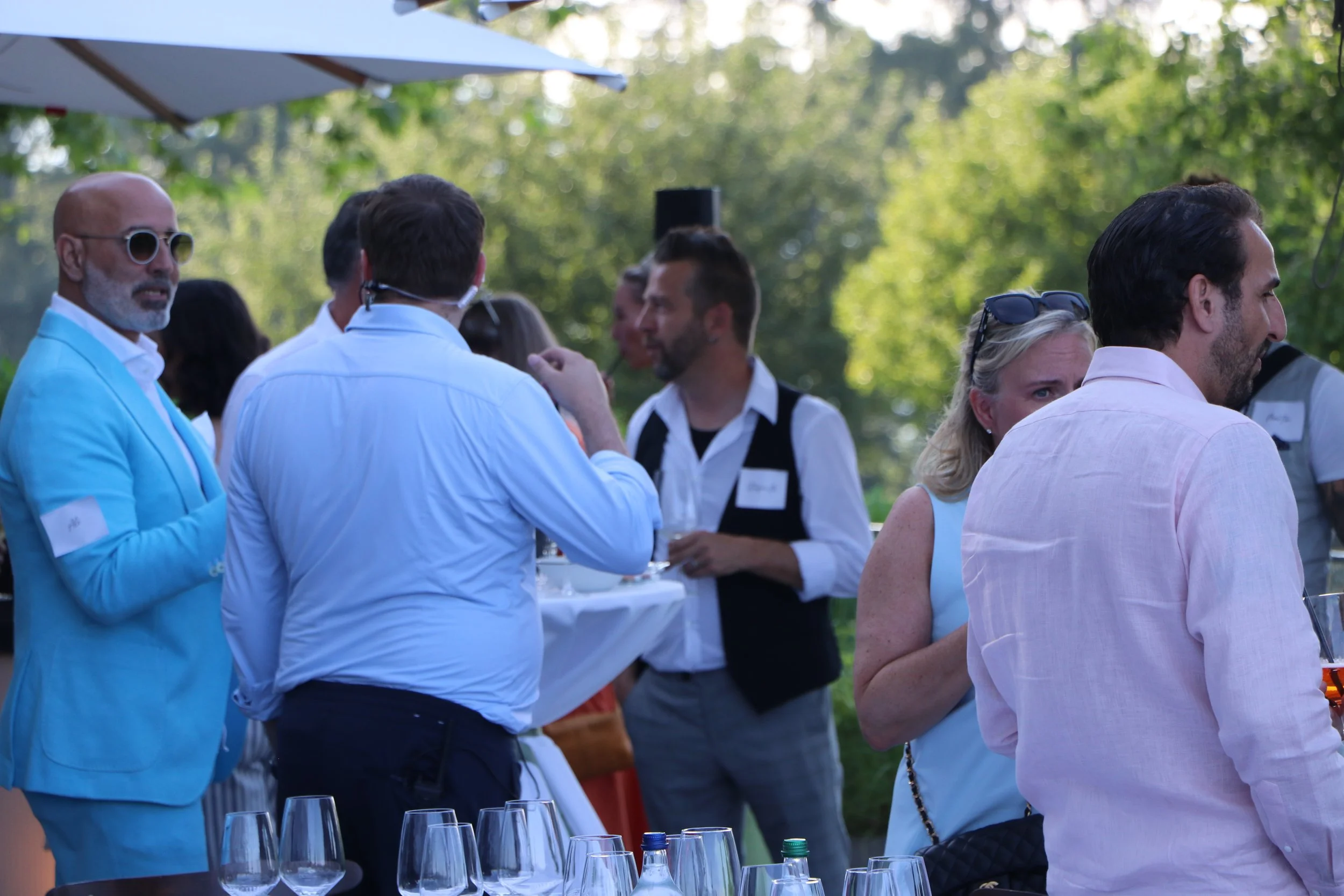 Menschen feiern im Freien bei einem Garten fest, mit Weingläsern auf einem Tisch im Vordergrund, und grünen Bäumen im Hintergrund.