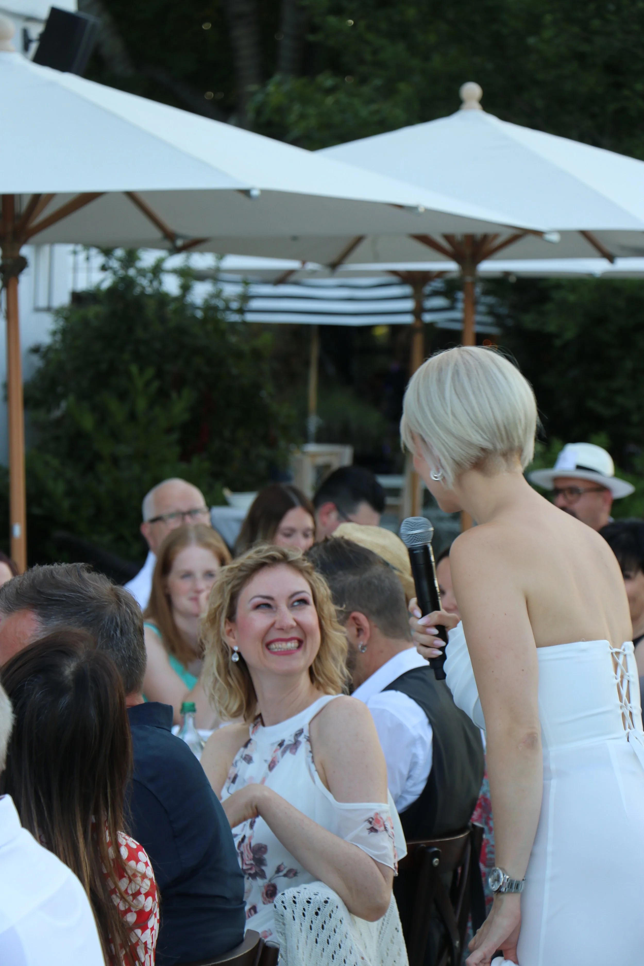 Eine Gruppe von Menschen bei einem Außenessen unter Sonnenschirmen, eine Frau mit kurzem blonden Haar hält ein Mikrofon und spricht zu einer lachenden Frau, die andere Menschen im Hintergrund beobachten.