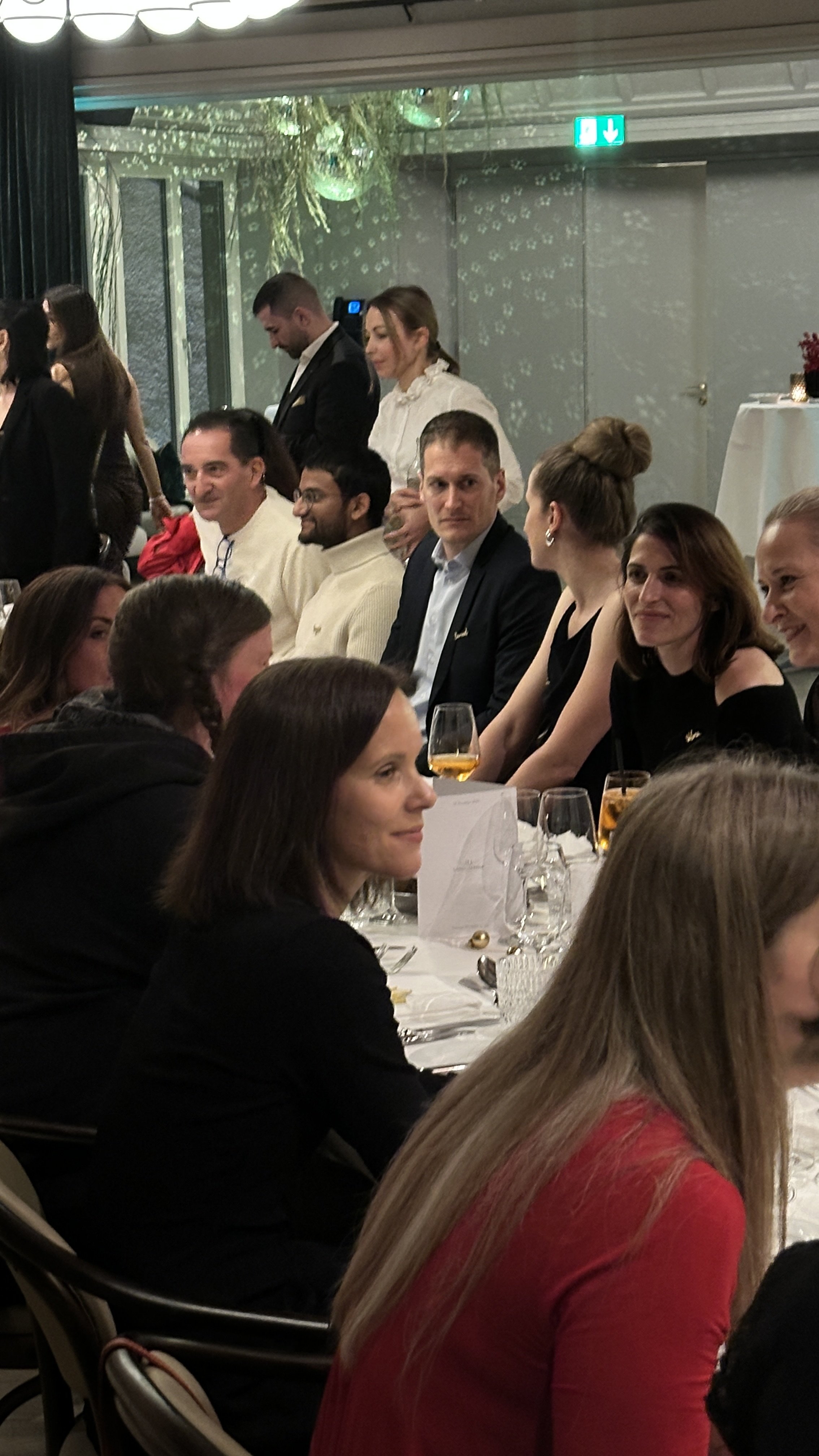 Menschen sitzen bei einer Veranstaltung an einem festlich gedeckten Tisch, einige trinken Getränke, im Hintergrund sind weitere Personen und Schattenprojektionen an der Wand zu sehen.