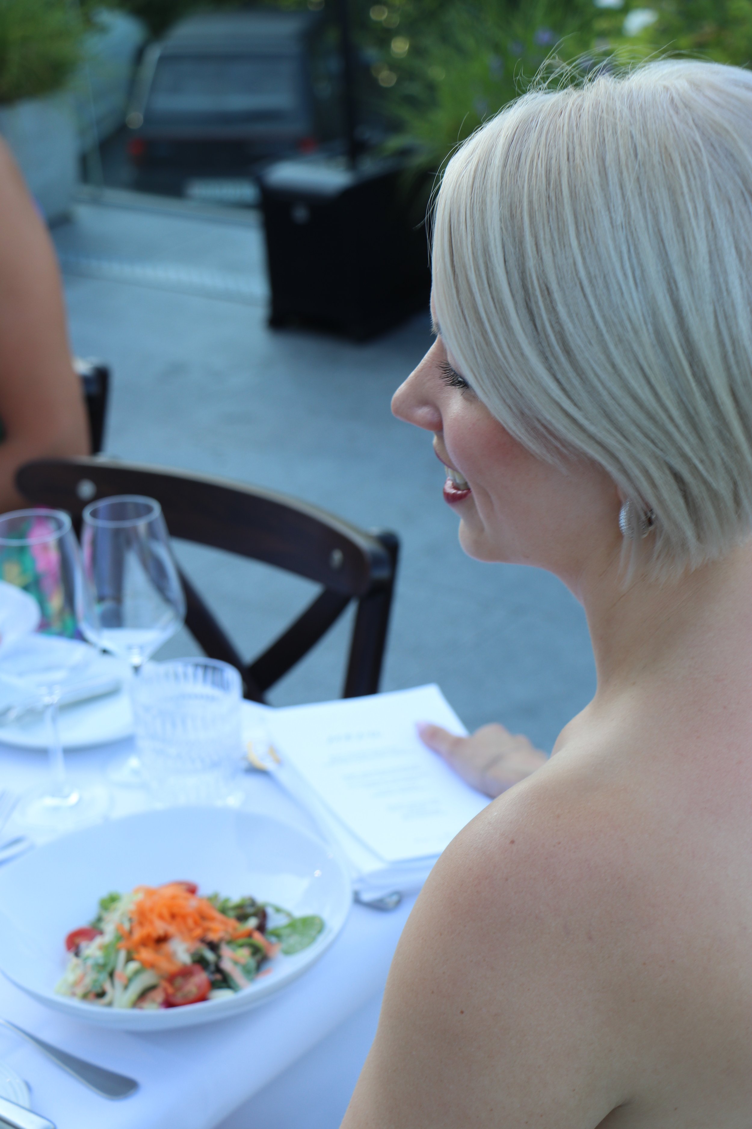 Eine blond geschminkte Frau sitzt bei einem Abendessen im Freien, vor einem Salat und Gläser auf einem Tisch. Sie liest ein Menü. Im Hintergrund ein Auto und grüne Pflanzen.