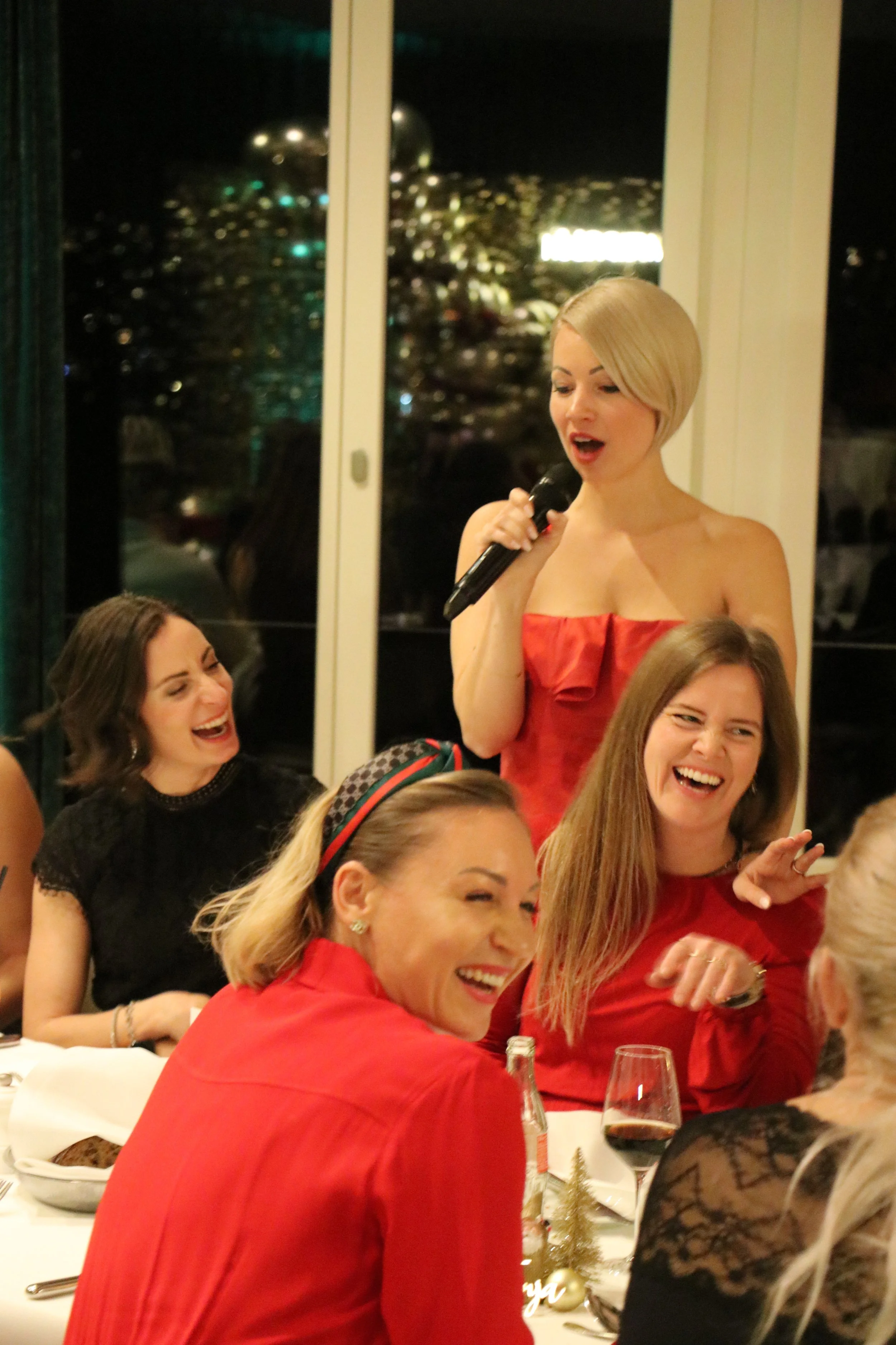 Eine Frau hält eine Mikrofon bei einer Feier, umgeben von lachenden und fröhlichen Gästen.